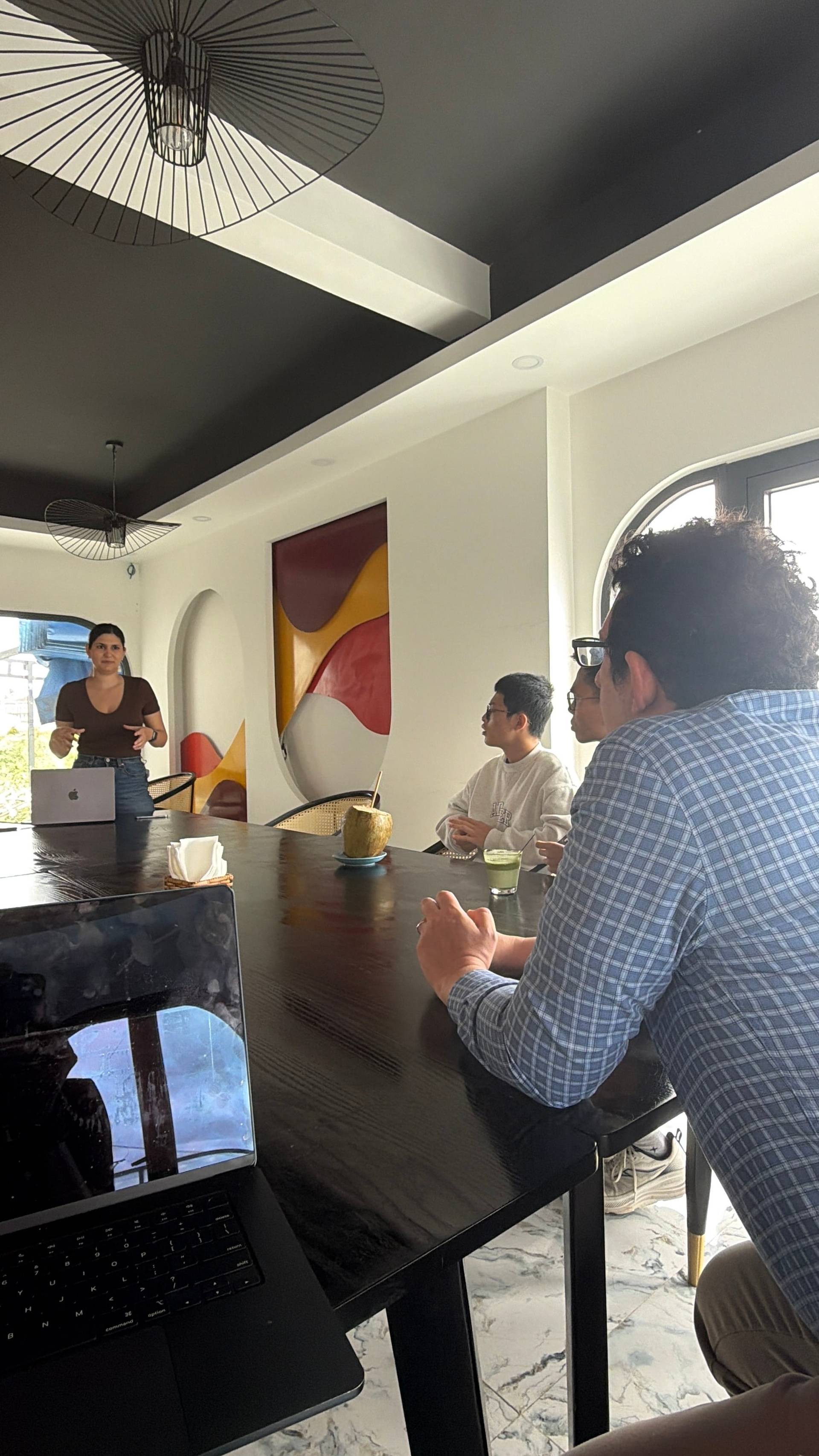 A group gathered in a modern Đà Lạt venue for a presentation or workshop, featuring contemporary design with abstract wall art and designer lighting, participants engaged in active listening and discussion around a sleek black table.