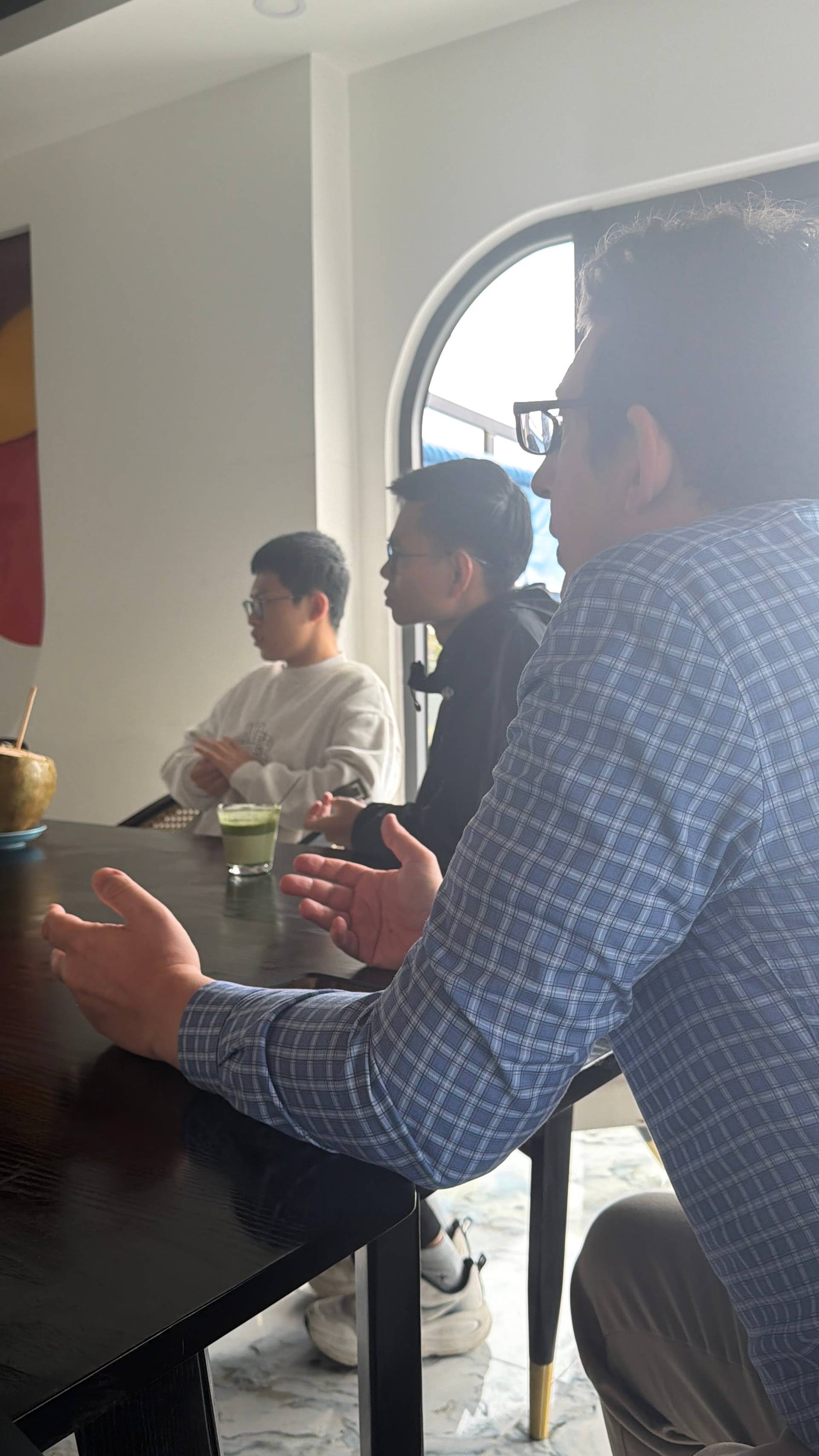A collaborative discussion unfolds around a table in a modern Đà Lạt event space, with participants engaged in conversation framed by large arched windows overlooking the landscape.