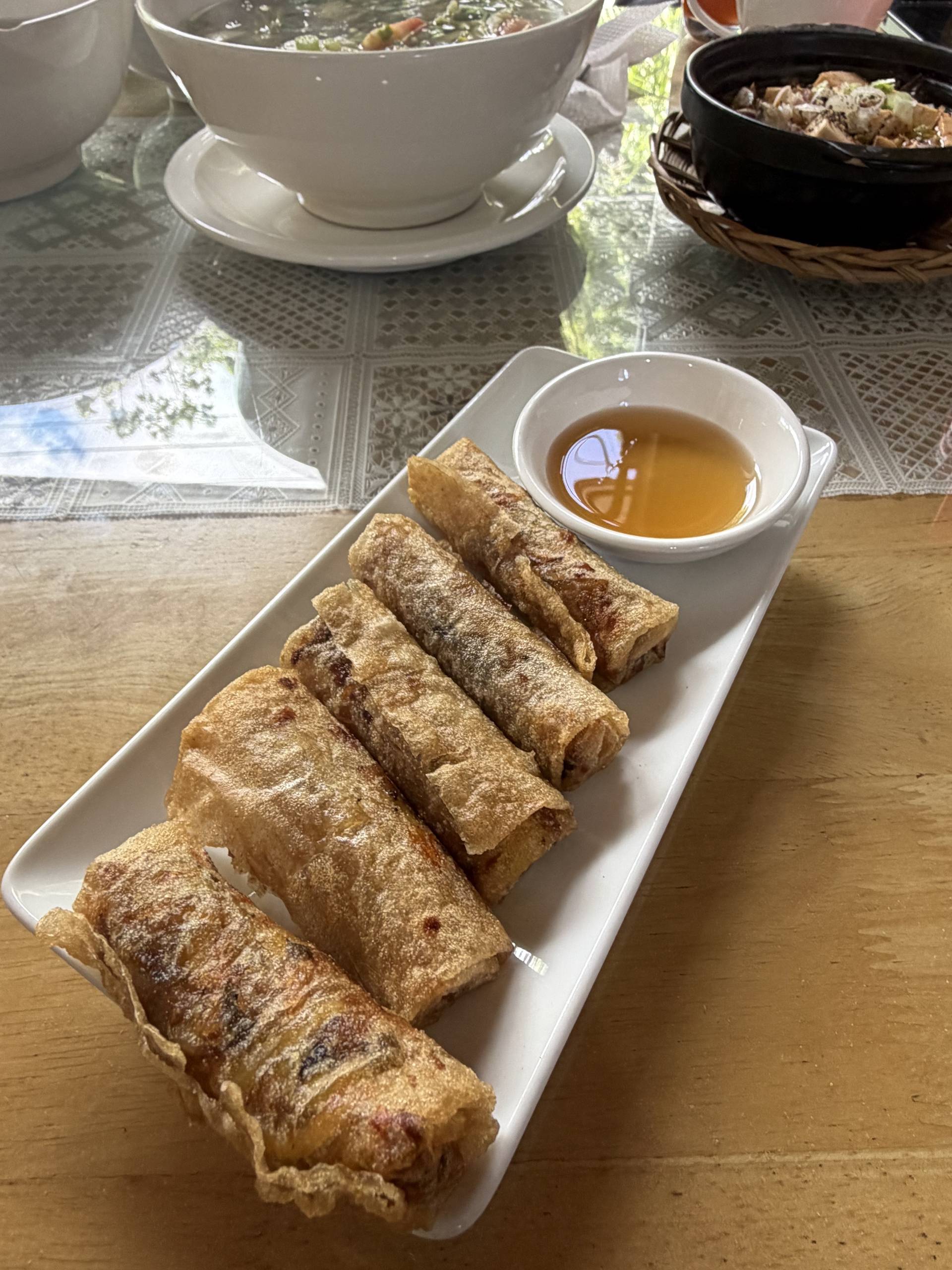 Golden fried spring rolls arranged on a white plate with aromatic dipping sauce, alongside a traditional noodle soup. Authentic Vietnamese culinary experience in Đà Lạt.