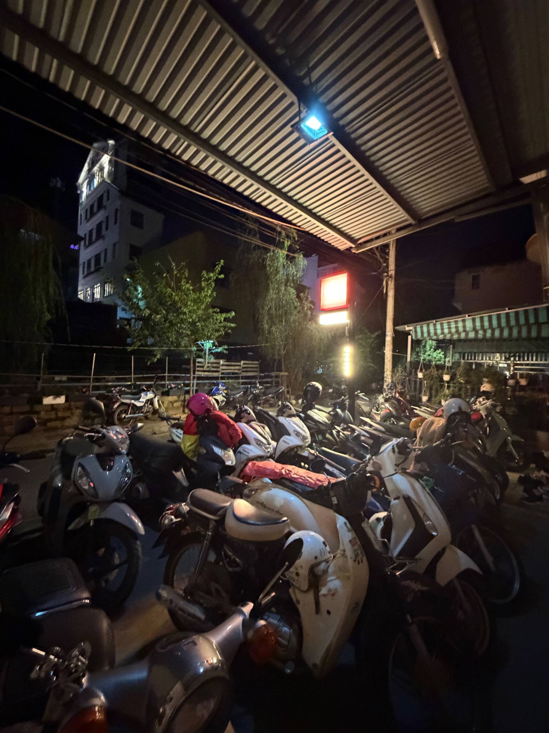 A bustling nighttime scene in Đà Lạt, Vietnam, with rows of parked motorcycles under a covered area illuminated by streetlights.