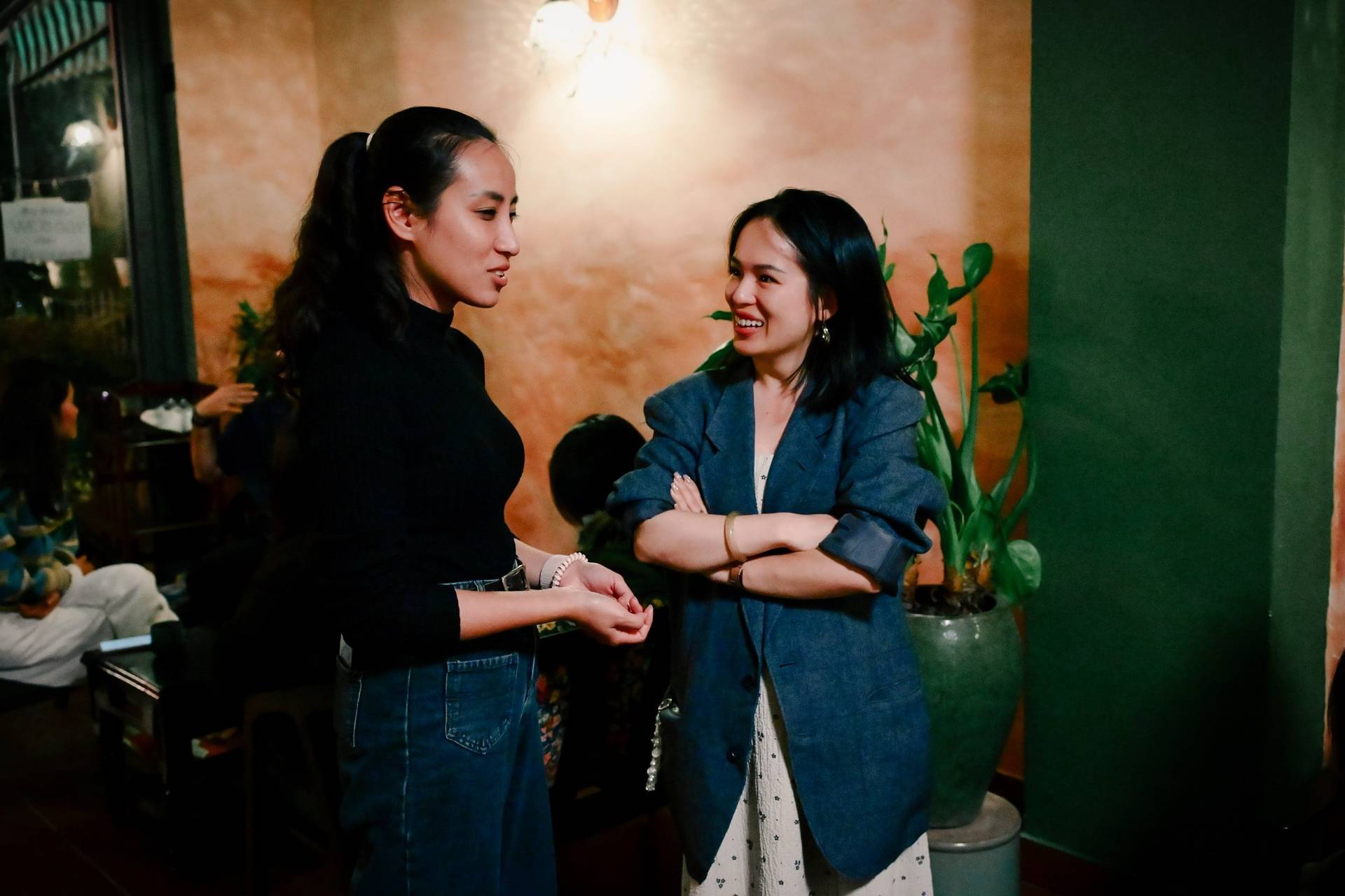 Two women engage in a lively conversation in a cozy café setting with warm lighting and greenery, reflecting a vibrant yet intimate atmosphere.