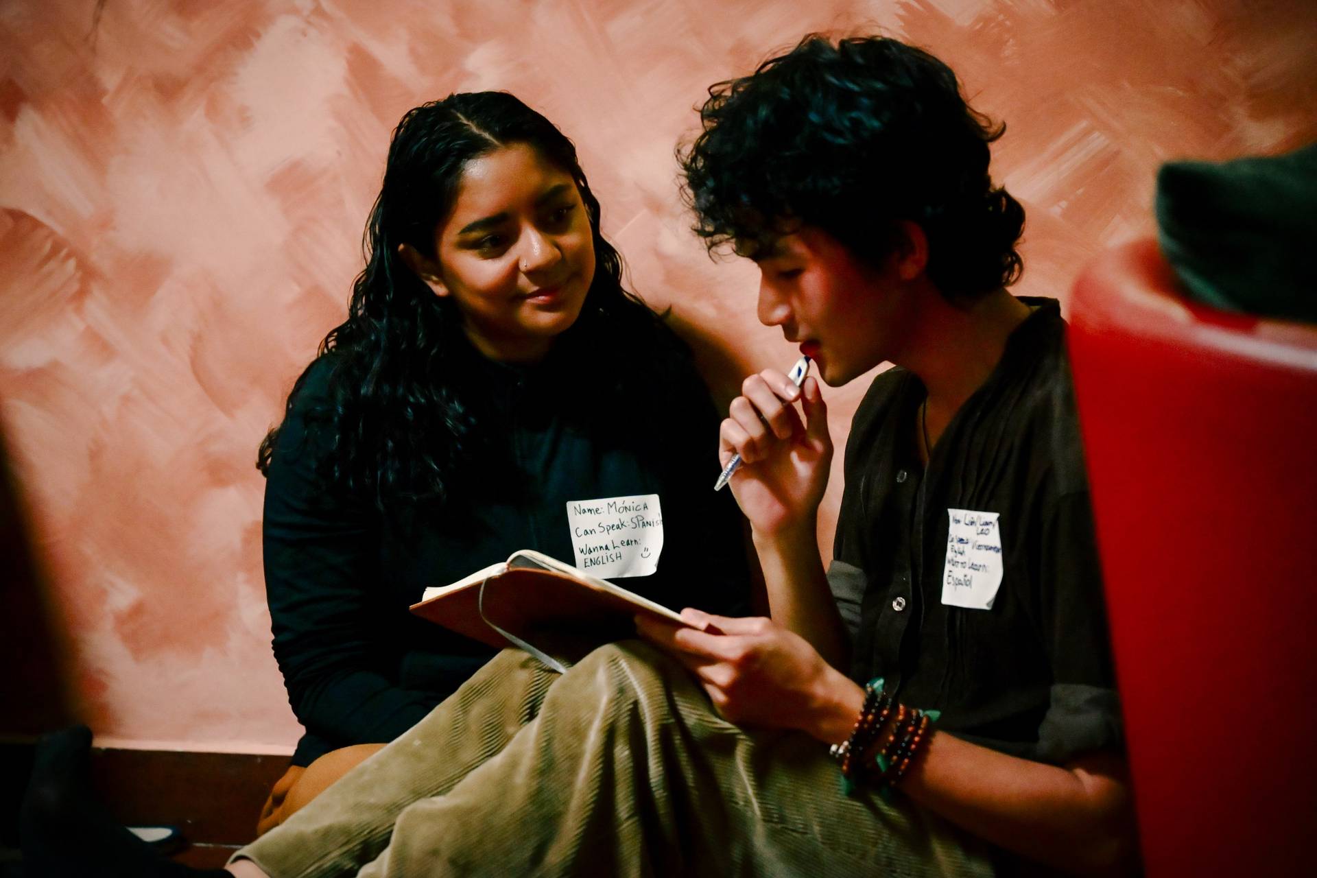 Two individuals engage in a conversation during a social gathering in Đà Lạt, Vietnam, with one person holding a notebook and pen, suggesting an exchange of ideas or learning session.