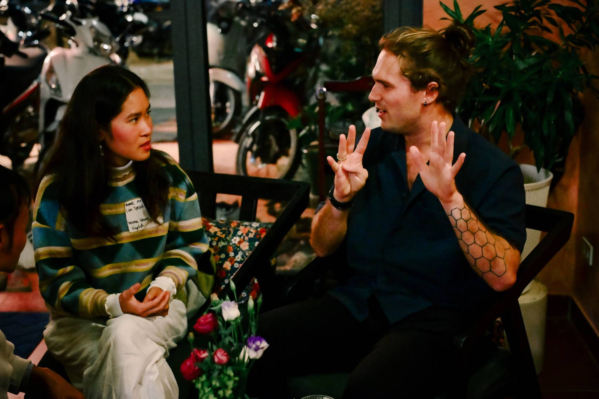 A lively conversation unfolds in a cozy café setting in Đà Lạt, Vietnam, with two individuals engaged in animated discussion over drinks and snacks.