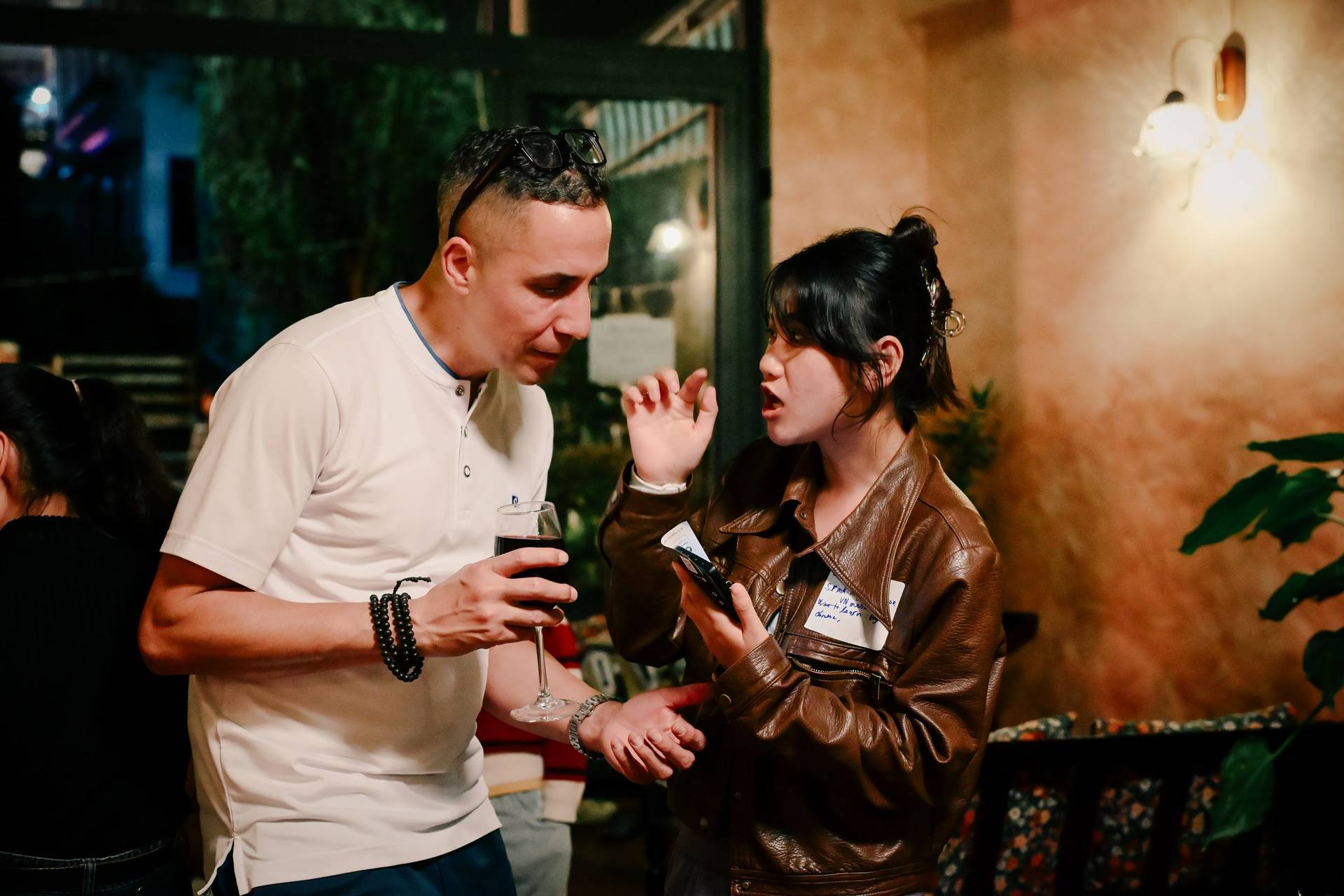 Two individuals engage in a lively conversation over drinks at a social gathering in Đà Lạt, Vietnam, surrounded by warm lighting and greenery.
