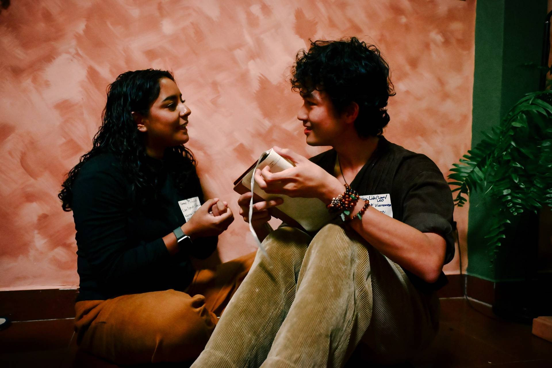 Two individuals engage in a casual conversation while seated on cushions against a textured wall backdrop in Đà Lạt, Vietnam.