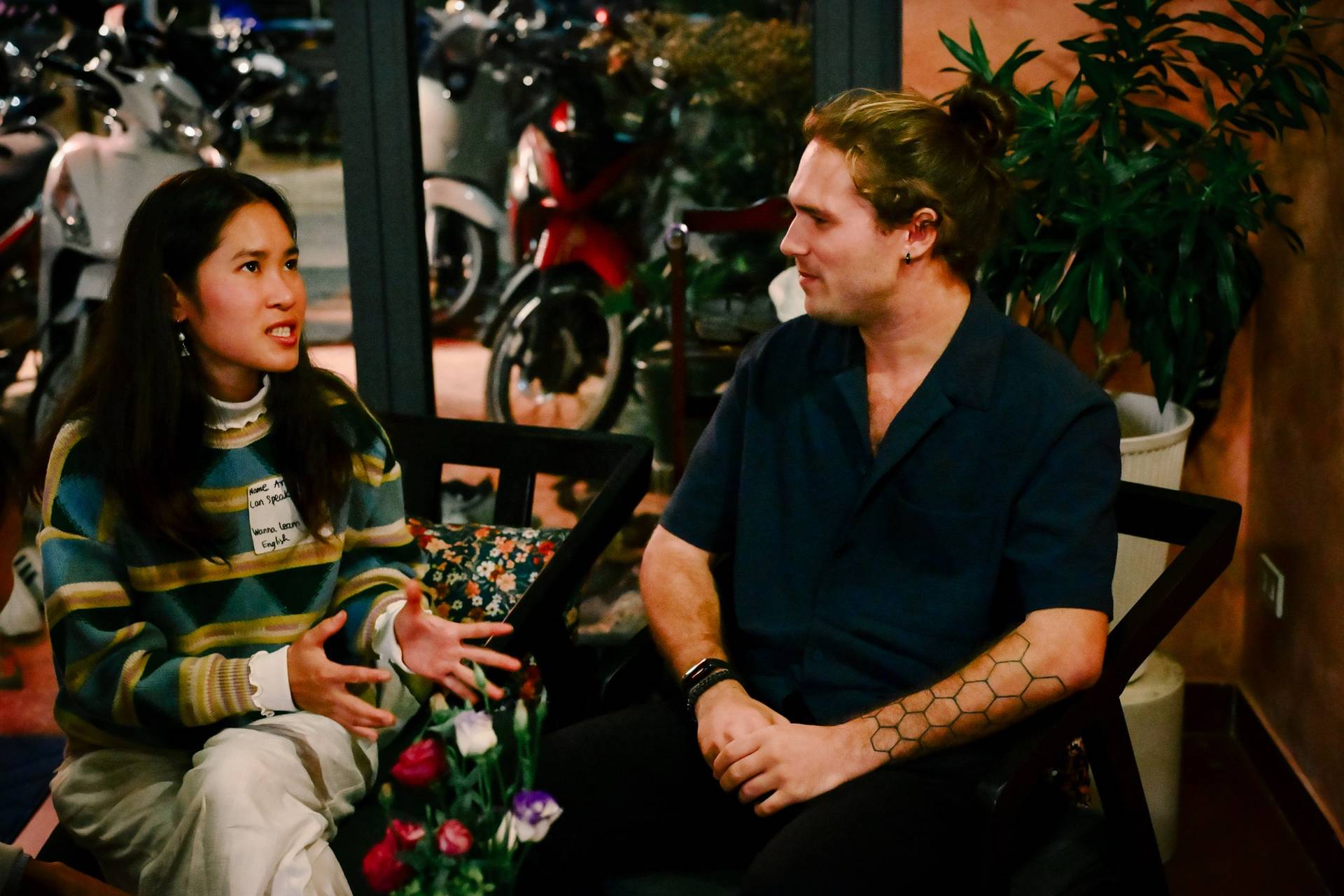 A casual conversation between two individuals in a cozy café setting with motorcycles visible through large windows, reflecting a relaxed and social atmosphere.