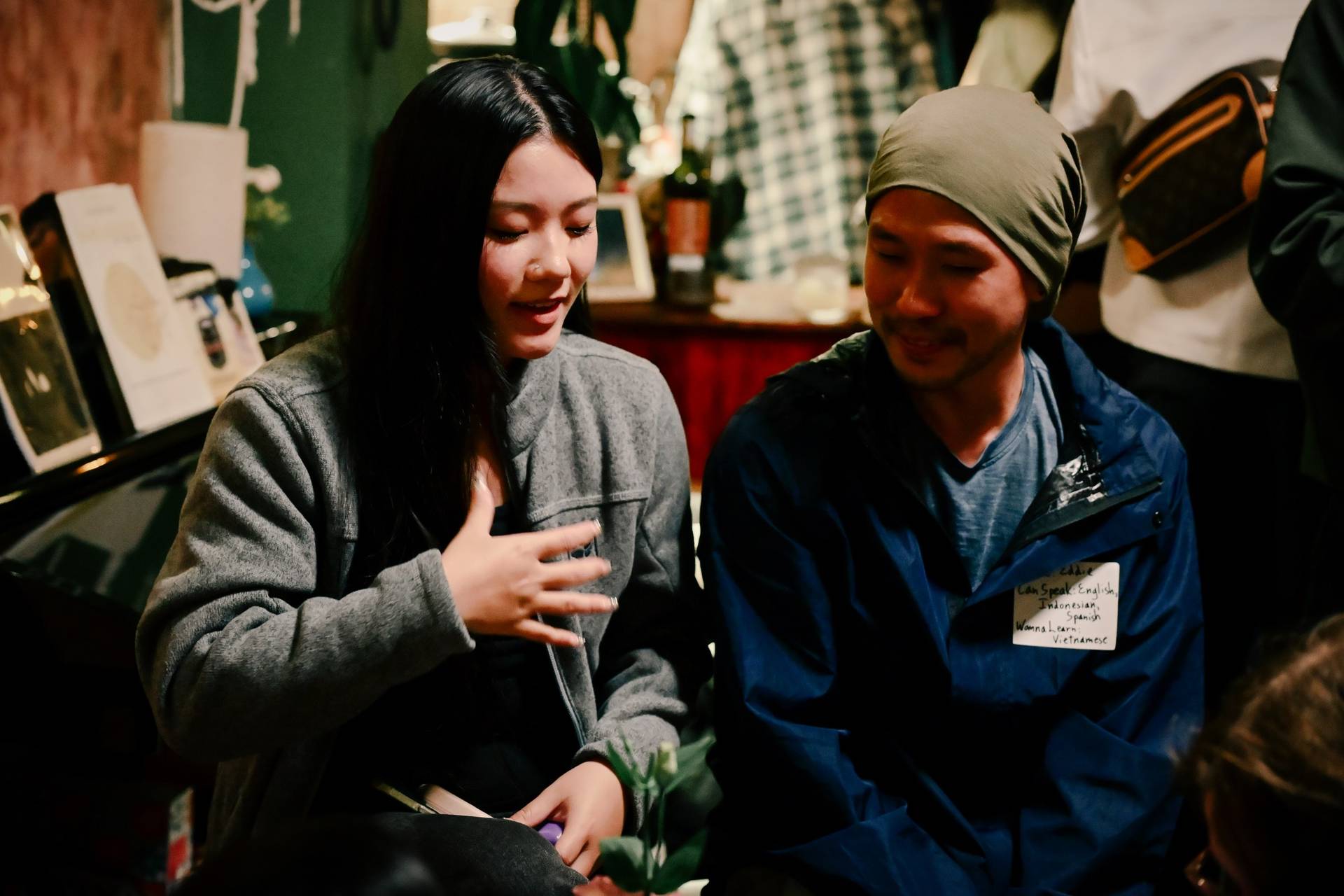 Two individuals engage in a lively conversation during a social gathering in Đà Lạt, Vietnam, surrounded by cozy decor and warm lighting.