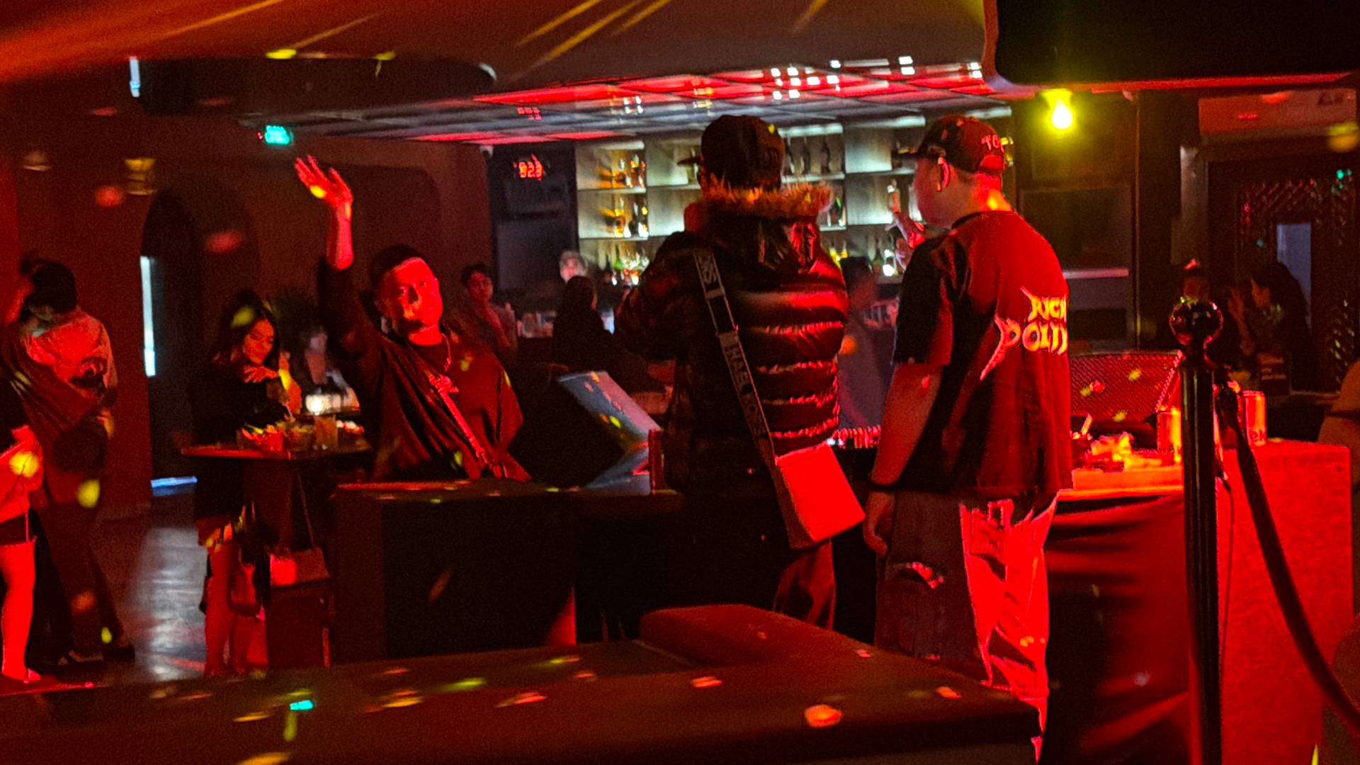 A crowded nightclub in Đà Lạt glows with intense red and yellow neon lighting, creating dramatic atmospheric effects. Guests gather throughout the venue while sophisticated bar shelving and overhead lighting design enhance the nightlife experience.