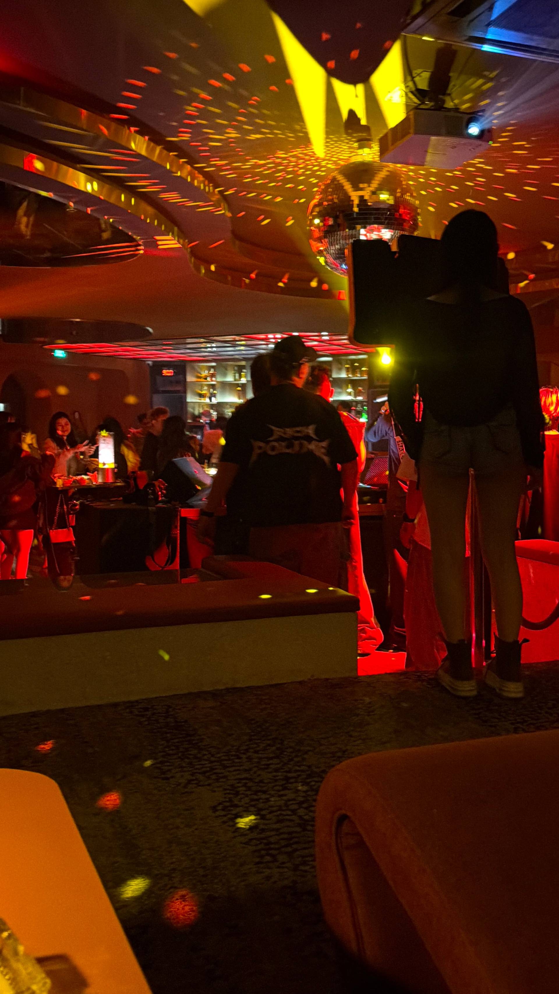 A high-energy nightclub scene in Đà Lạt featuring an impressive overhead lighting installation with yellow geometric shapes and red ambient lighting. Patrons mingle across the illuminated dance floor under the spectacular ceiling design.