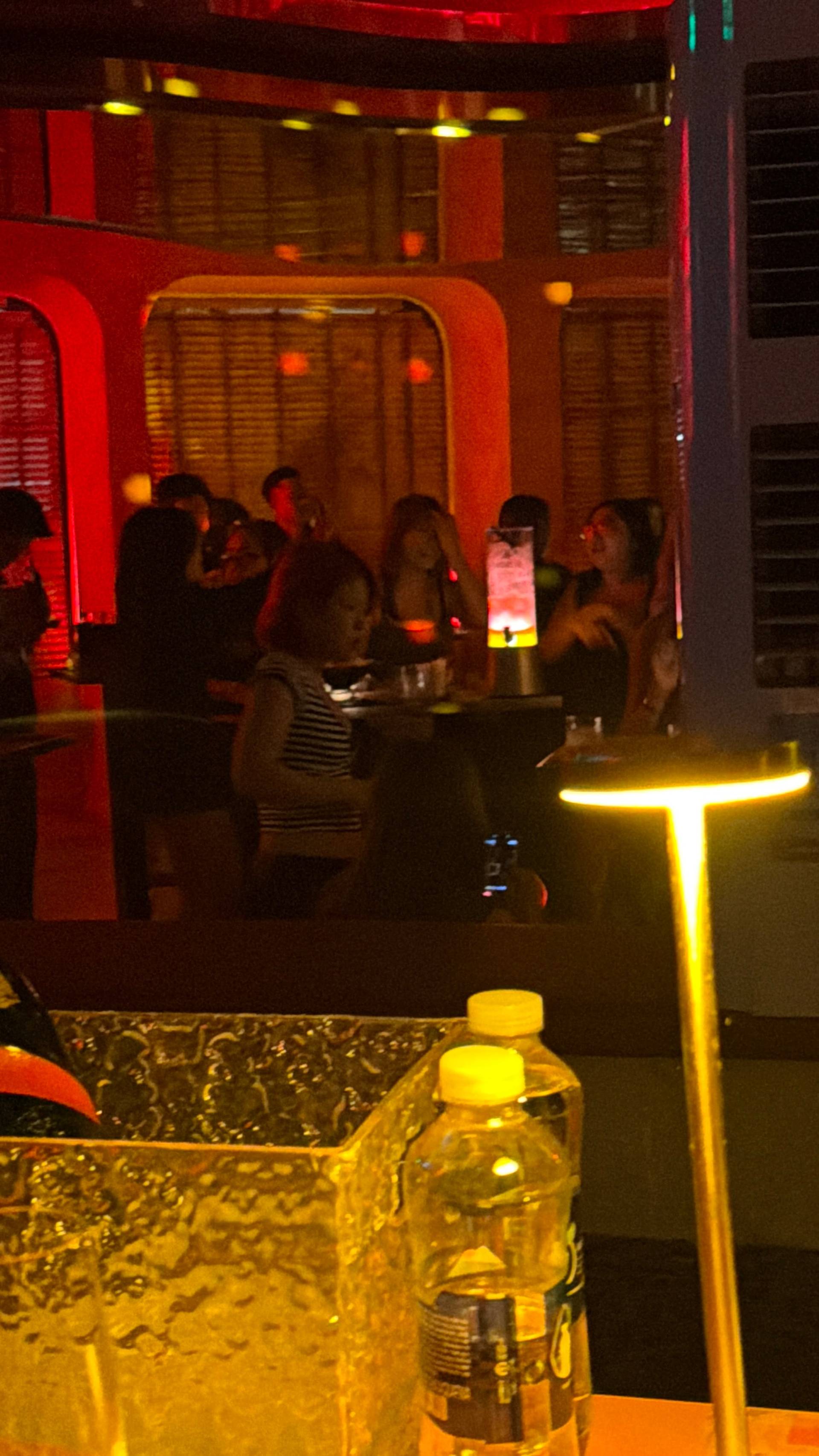 An intimate indoor lounge gathering in Đà Lạt with neon-accented red and yellow lighting, where guests congregate near a window seating area with decorative water bottles displayed on tables.