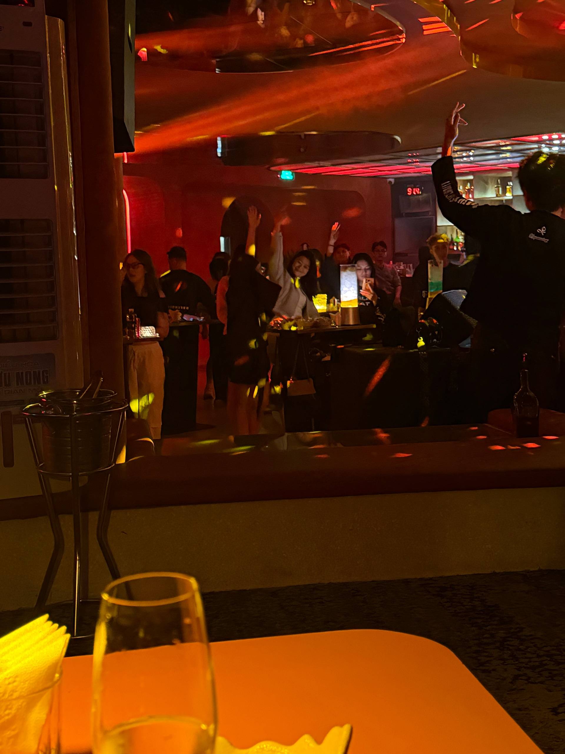 Guests celebrate at a modern nightclub lounge in Đà Lạt, with someone raising their hand in joy amid red and yellow atmospheric lighting. Glasses and bottles sit in the foreground as a group enjoys the energetic evening.