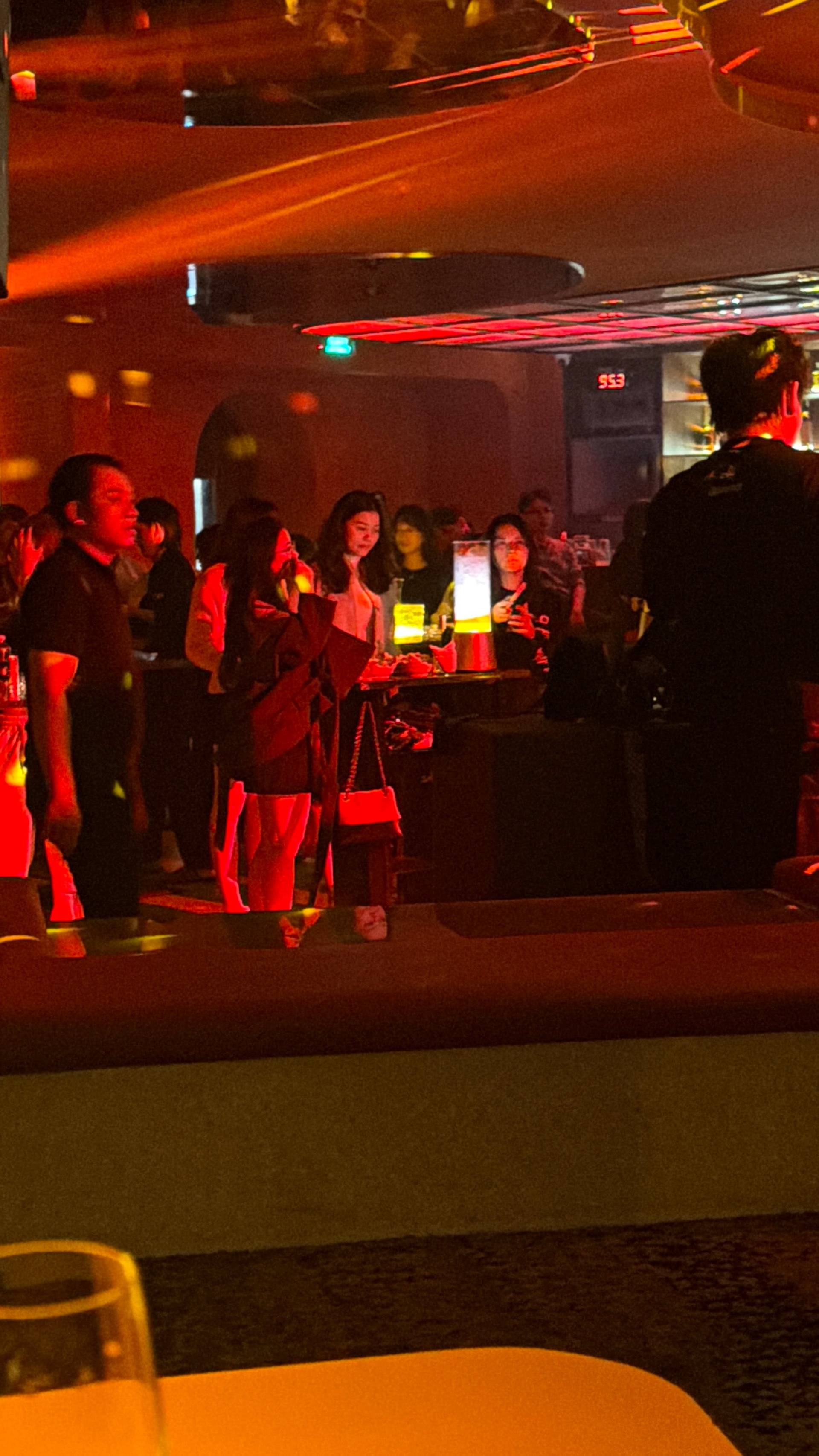 A group gathers inside a Đà Lạt nightclub under intense red and orange lighting, with some patrons holding phones and drinks while socializing. The sophisticated lighting and bar setup create an upscale nightlife experience.