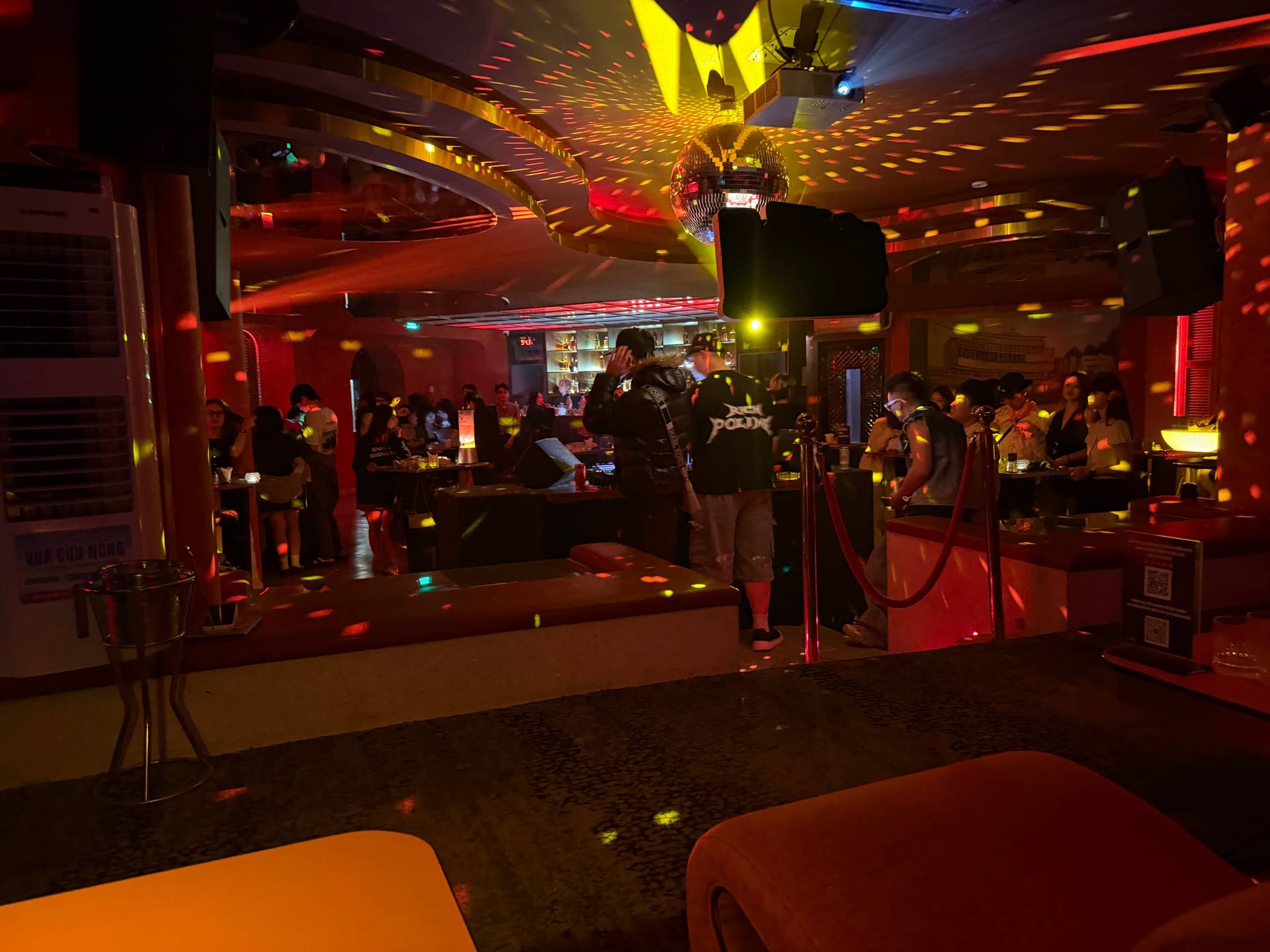 An intimate nightclub lounge in Đà Lạt showcases dramatic red and golden neon lighting with guests mingling at tables and the bar visible in the background. The layered lighting design and modern furnishings create a sophisticated evening atmosphere.