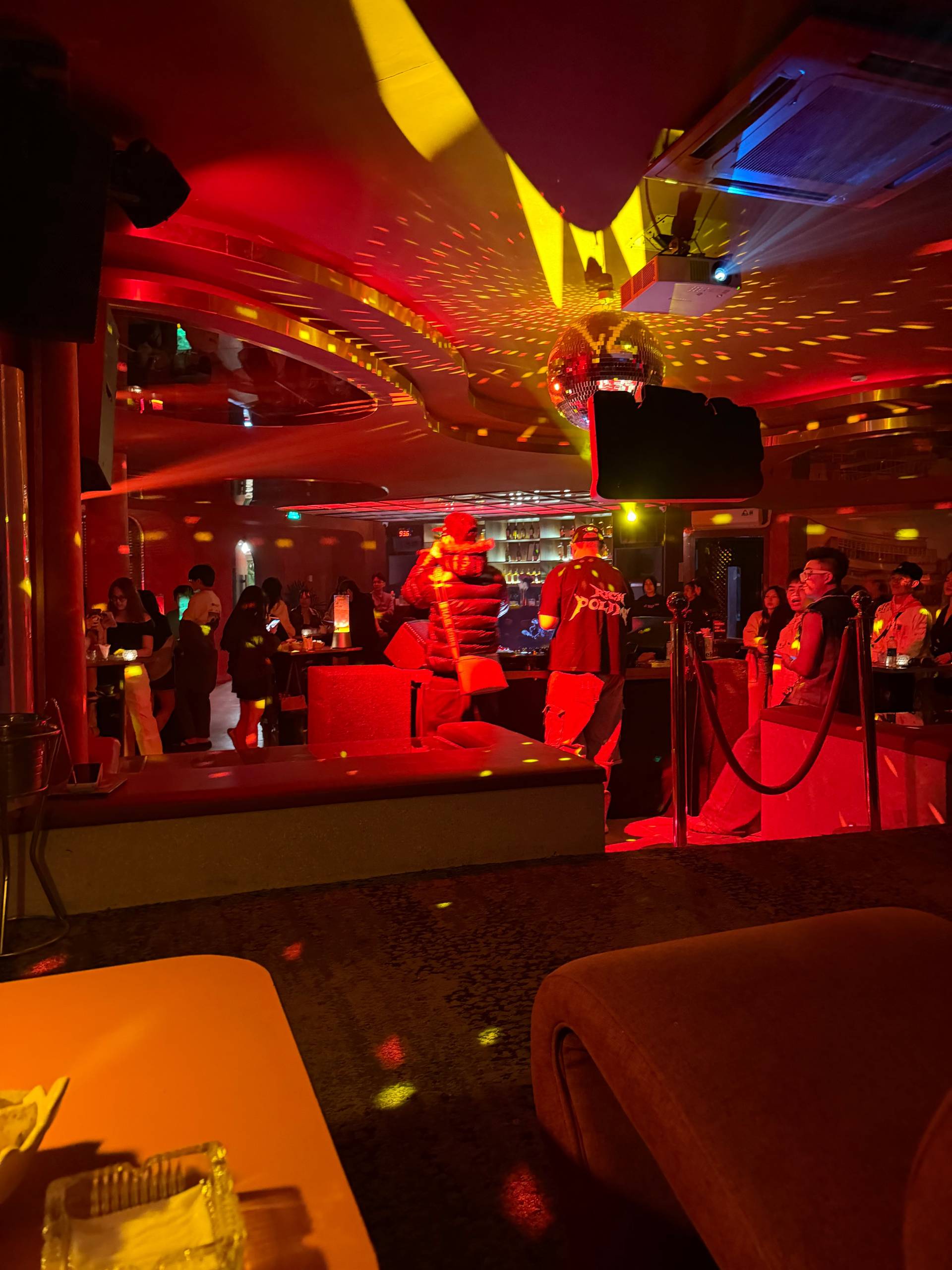 Lively nightclub interior in Đà Lạt bathed in vibrant red and golden neon lighting, with groups gathering at lounge areas and the bar. Geometric ceiling lights and dramatic ambient design create an energetic backdrop for evening socializing.