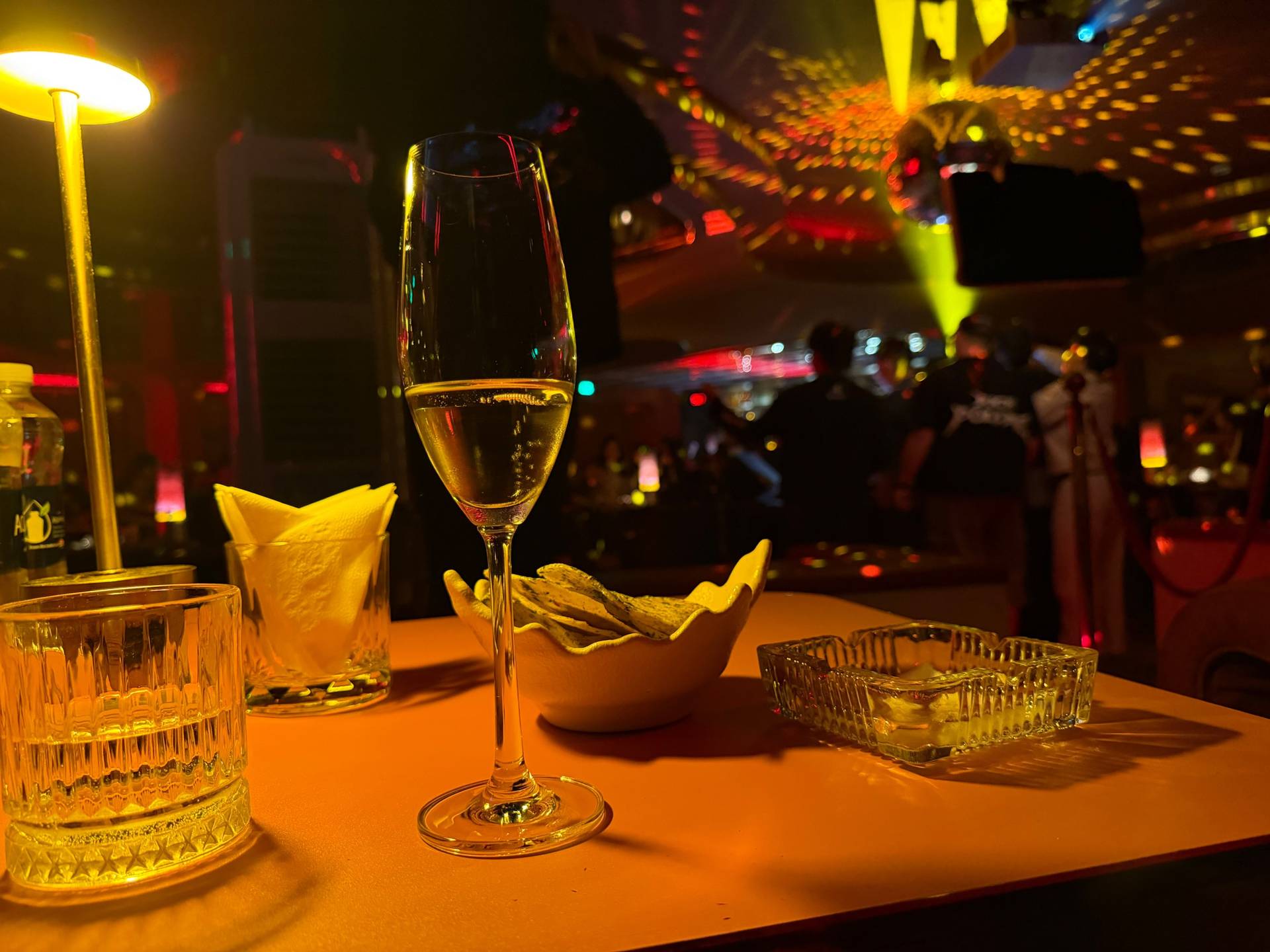 Elegant cocktails and appetizers at a vibrant nightclub in Đà Lạt, illuminated by warm golden and red neon lighting. Champagne glass, whiskey drink, and gourmet snacks set the tone for an upscale evening of celebration and socializing.