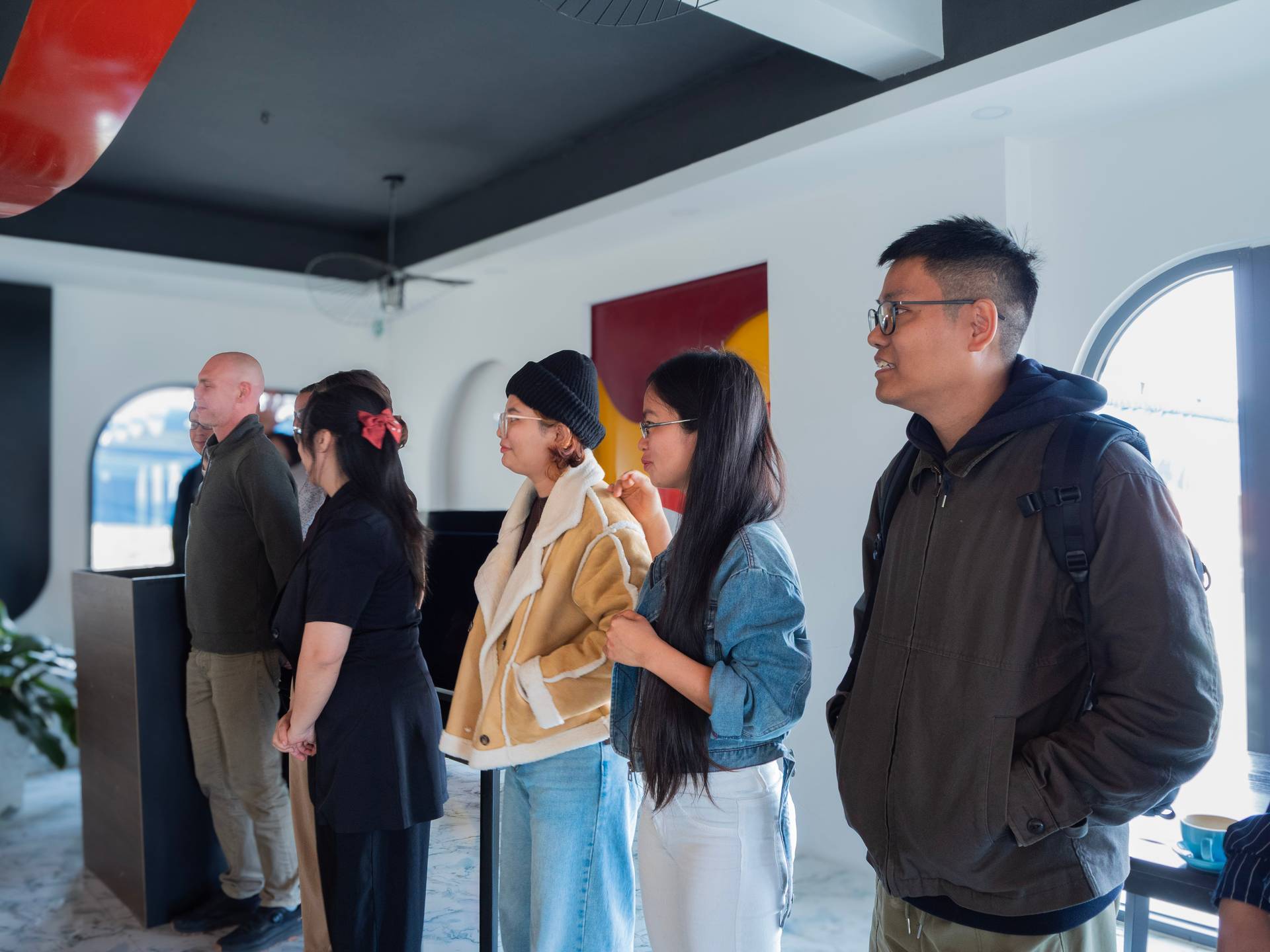 A diverse group of hackathon participants mingles in the modern Dalat Build Week venue, featuring contemporary architecture with red and yellow accent panels.