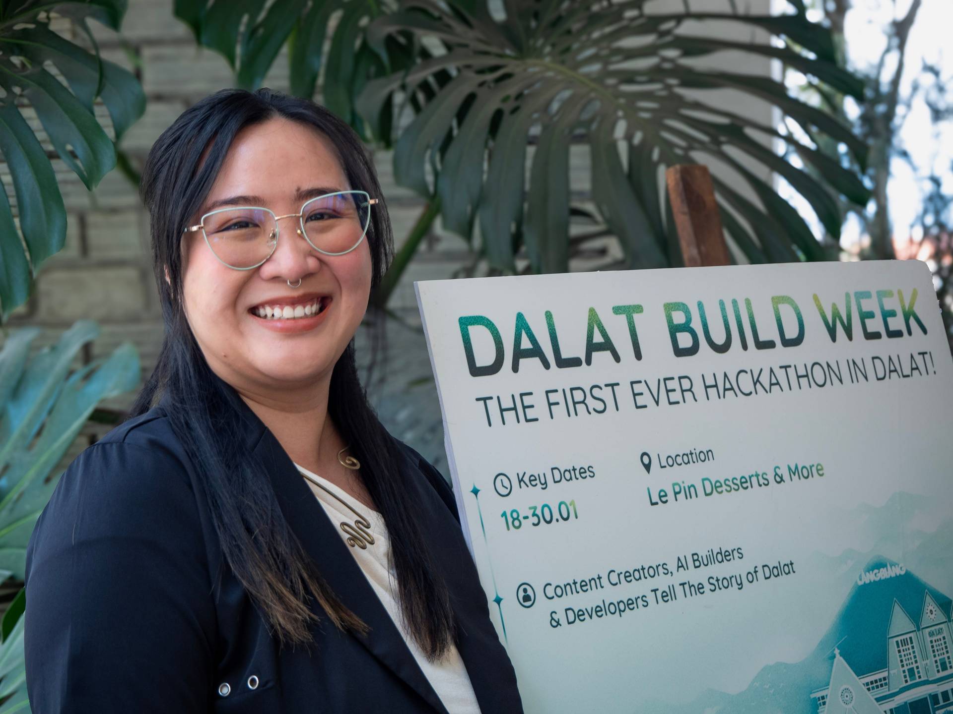 Close-up portrait of a Dalat Build Week participant smiling next to the event banner, with tropical plants creating a natural frame in this vibrant hackathon venue.
