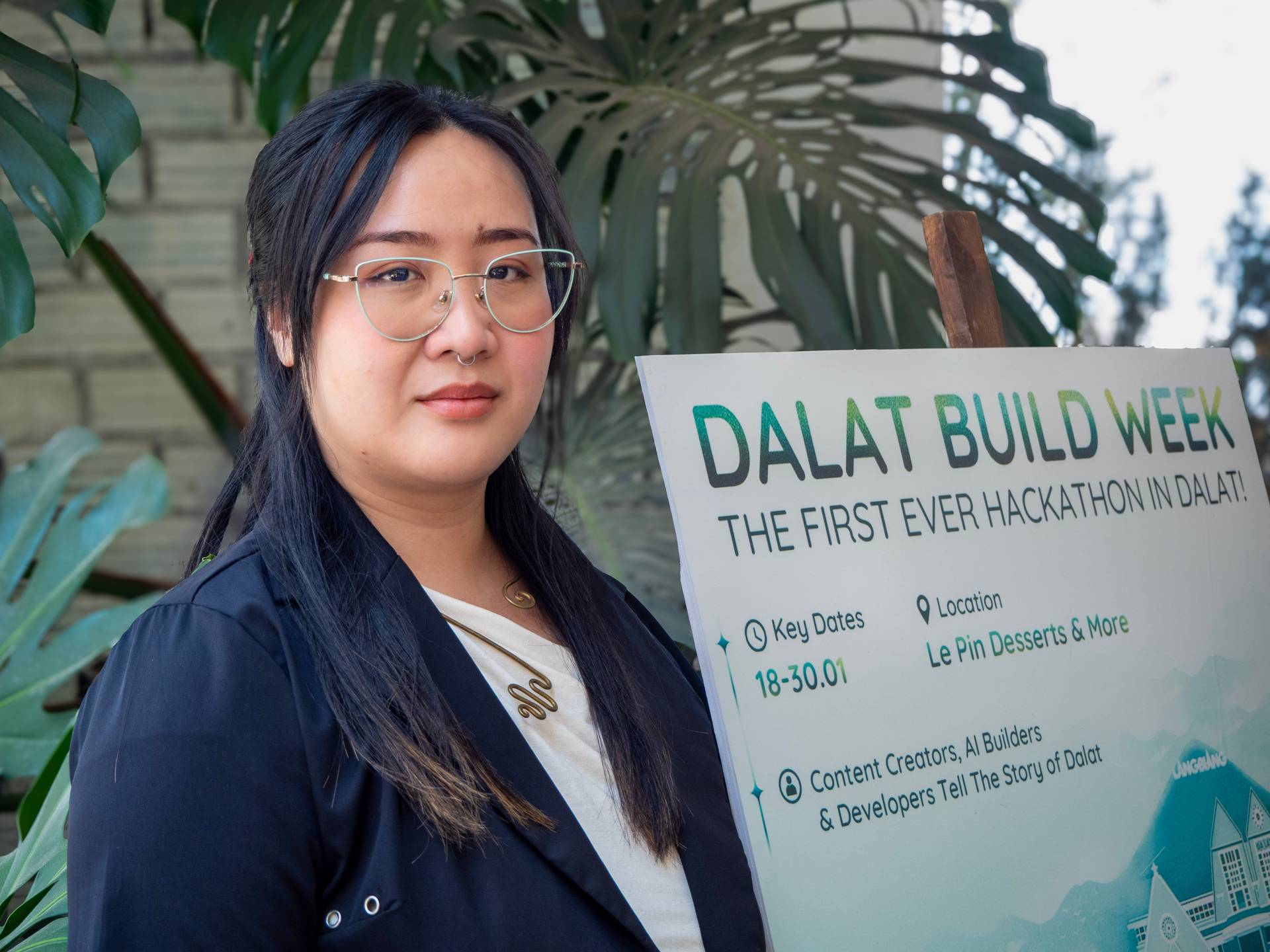 An event participant poses with the Dalat Build Week hackathon banner in a plant-filled venue, showcasing the first-ever hackathon initiative in Đà Lạt.