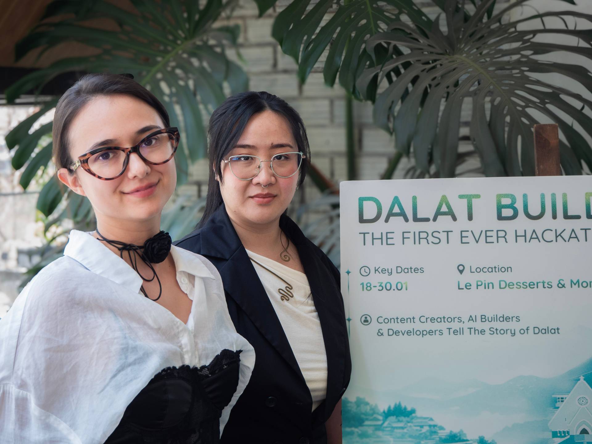 Two participants pose with the official Dalat Build hackathon event banner, surrounded by tropical foliage at Le Pin Desserts & More venue.