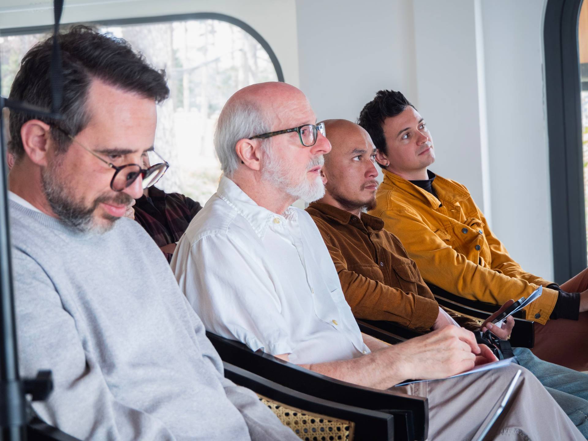Event attendees seated and facing toward a presentation, with some holding devices and phones, in a bright minimalist conference space.