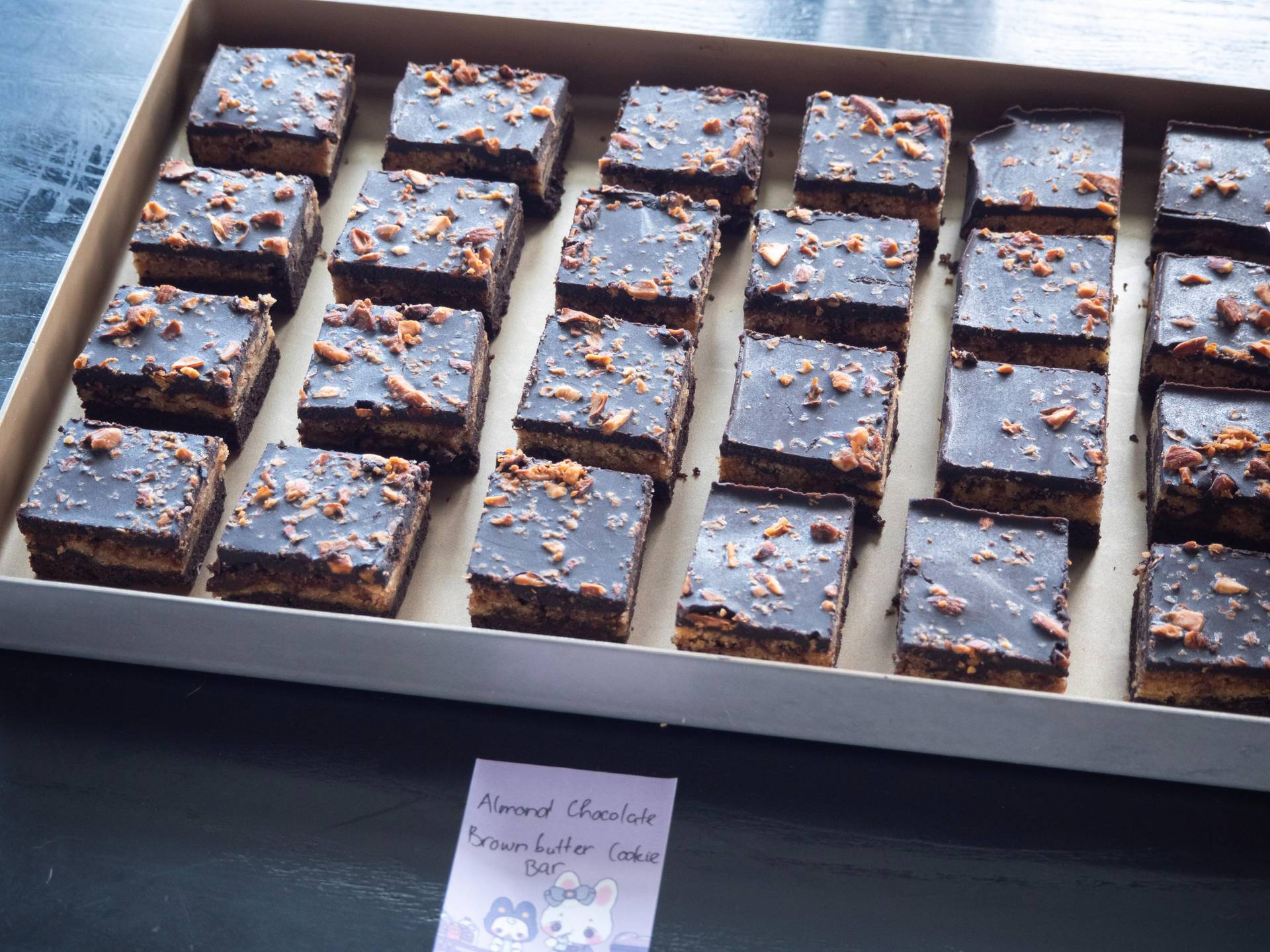 Homemade or artisan chocolate almond brownie bars with rich chocolate icing and toasted almond toppings. Displayed in a white presentation box with handwritten label, typical of Đà Lạt's creative food culture.