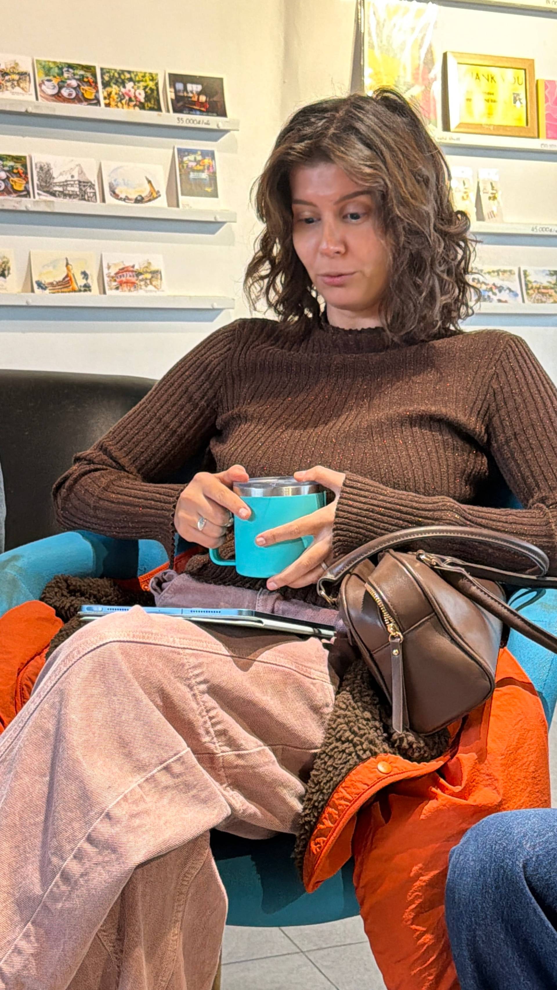 Cozy café gallery moment in Đà Lạt with turquoise mug and colorful seating. Framed artwork and Vietnamese postcards adorn modern display shelves behind. A warm, intimate gathering space.