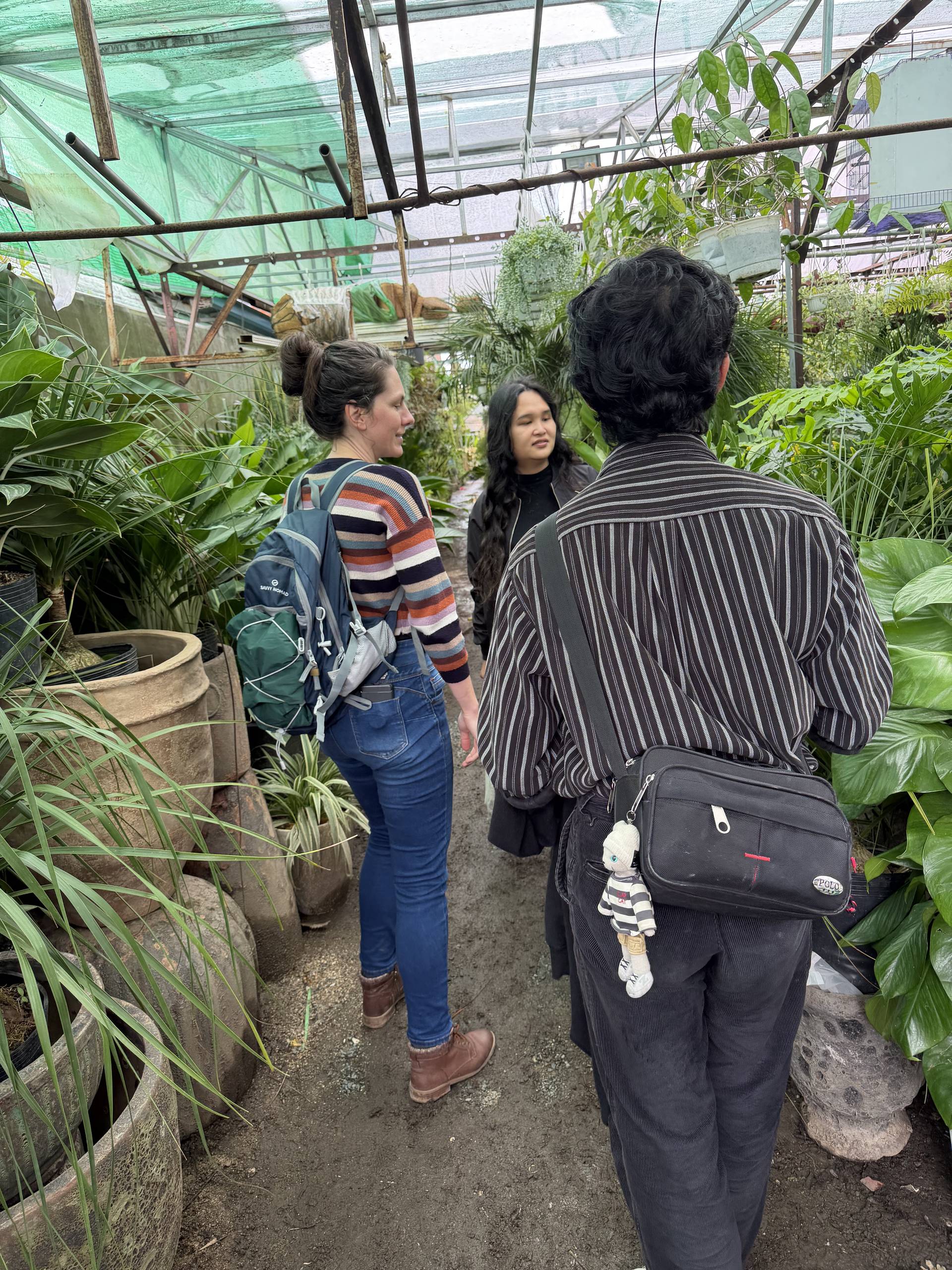 A group of three individuals is engaged in a conversation within a lush greenhouse filled with various plants and pots, suggesting an exploration or educational activity.