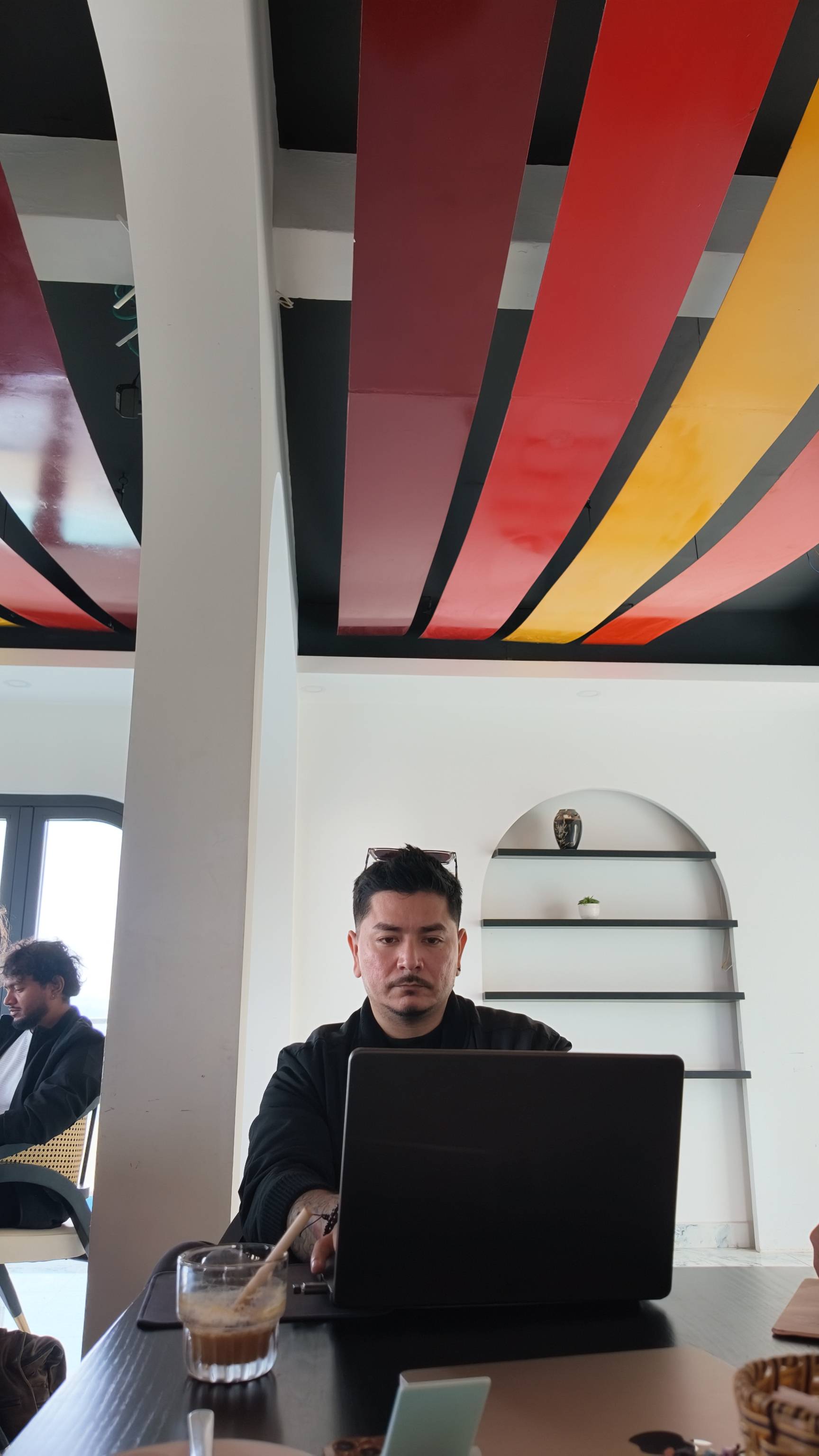 A man is focused on his laptop in a vibrant café setting with colorful ceiling decor and modern furnishings.