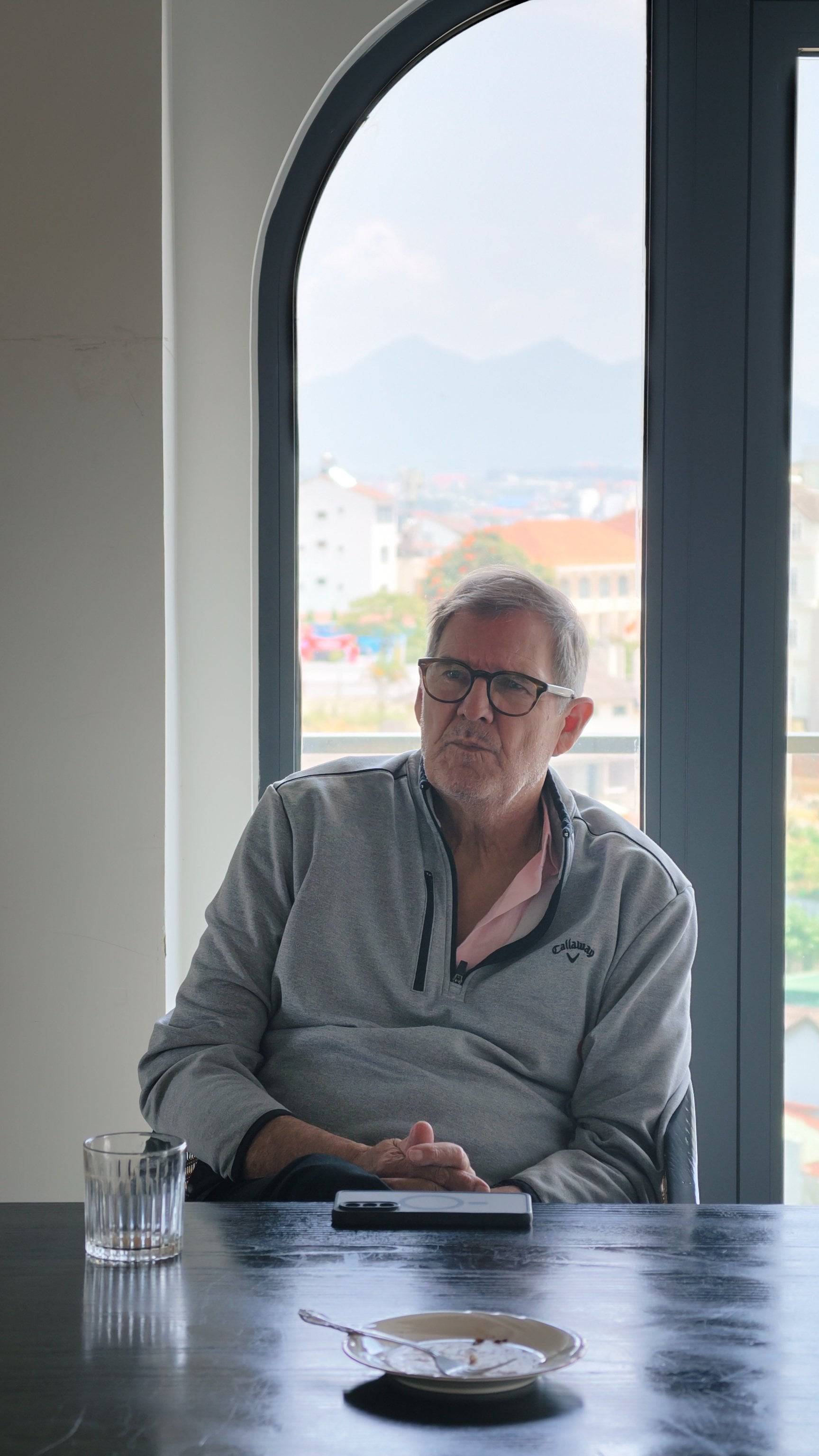A serene moment in Đà Lạt, Vietnam, where a person sits by a window overlooking scenic views of the city and mountains, enjoying a quiet meal with a glass of water.