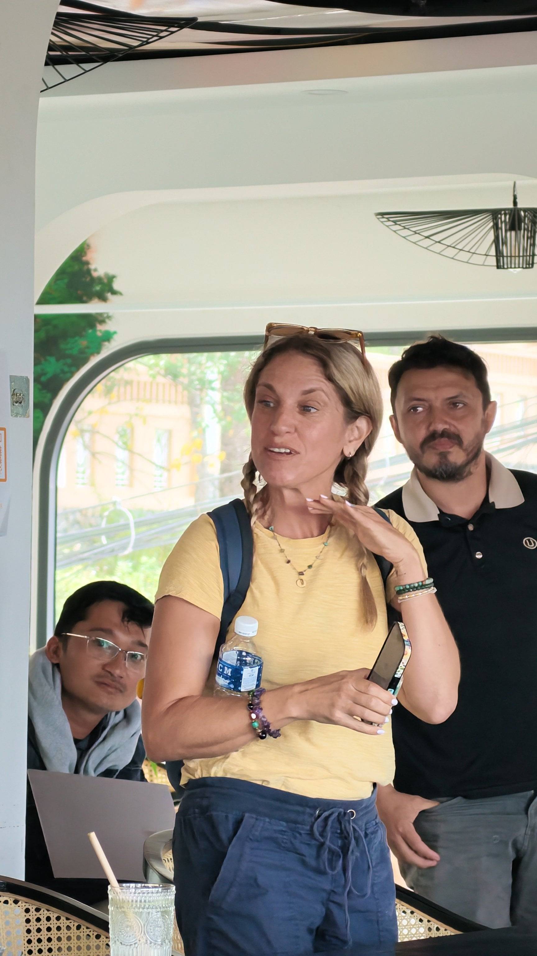 A woman in a yellow shirt stands confidently during what appears to be a presentation or discussion in a modern Vietnamese setting with natural light and greenery visible through large windows.