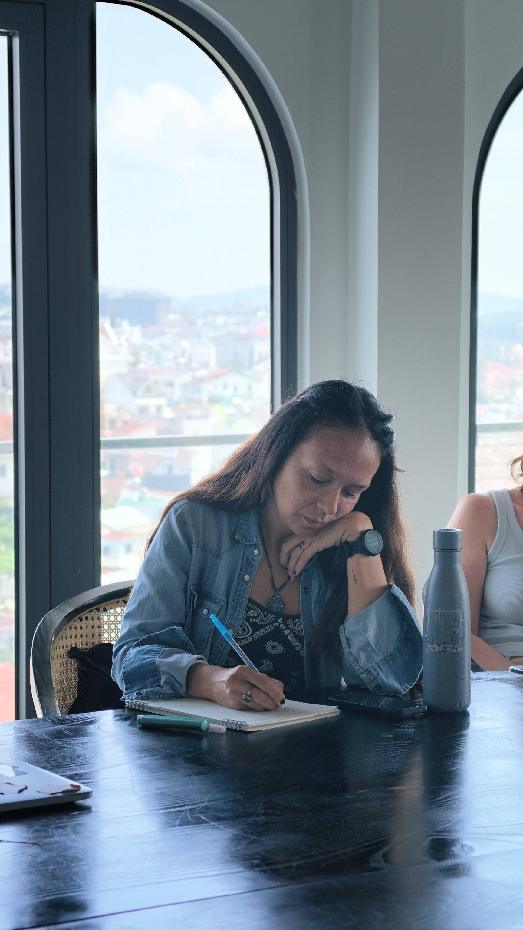 A woman is focused on writing in a notebook while seated by a large window with a scenic view of Đà Lạt's cityscape.