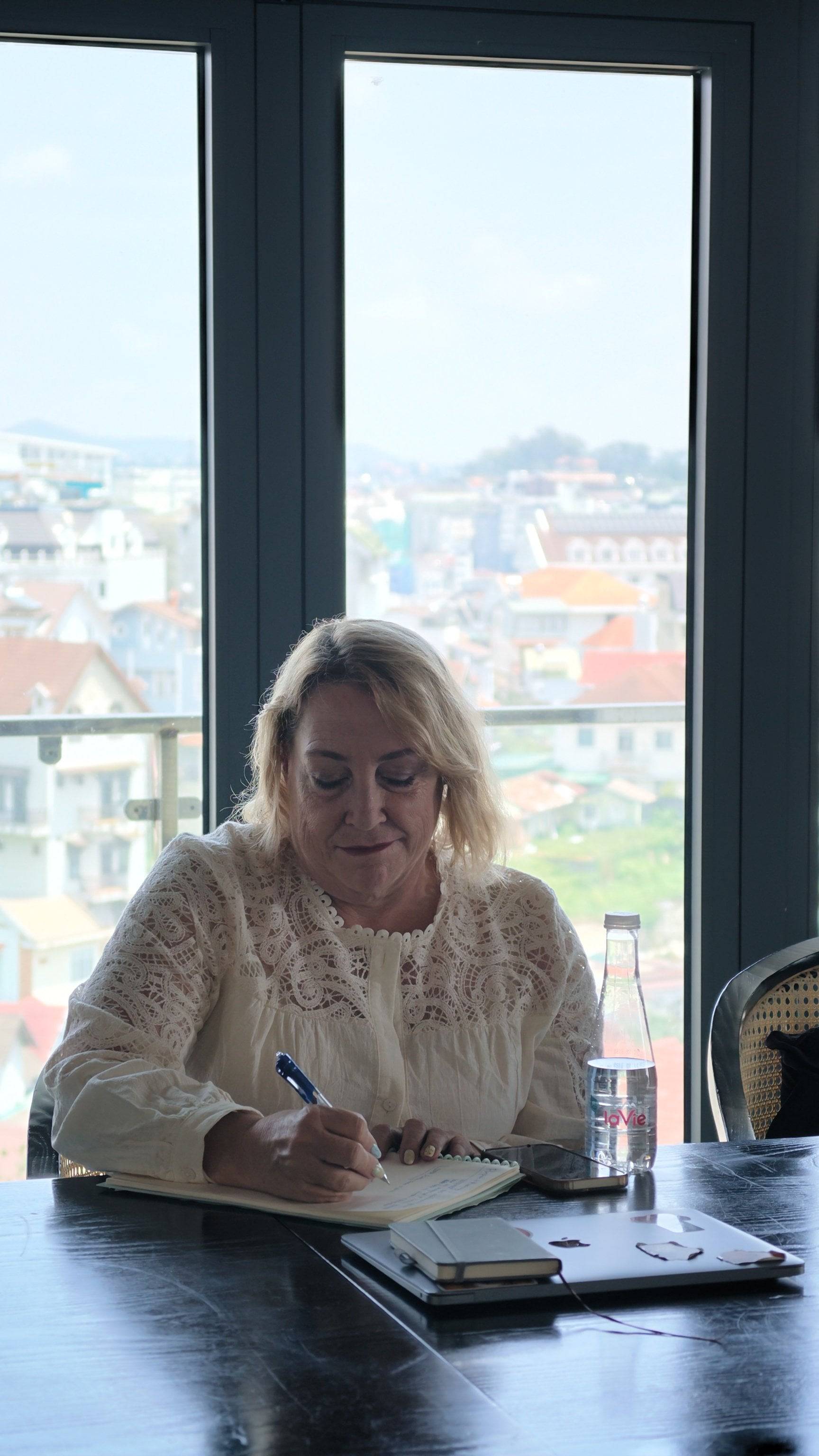 A woman is writing in a notebook with a scenic view of Đà Lạt's cityscape visible through large windows behind her.