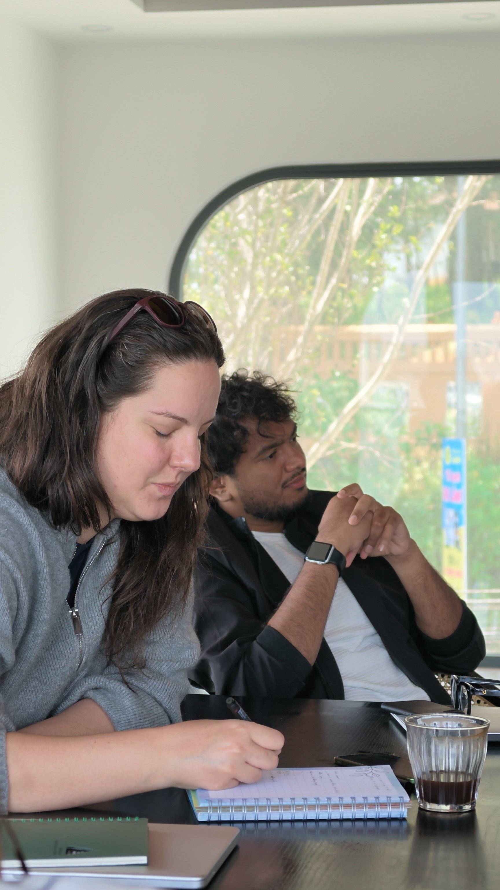 Two individuals are engaged in a focused discussion or meeting at a café in Đà Lạt, Vietnam, with natural light streaming through large windows and greenery visible outside.