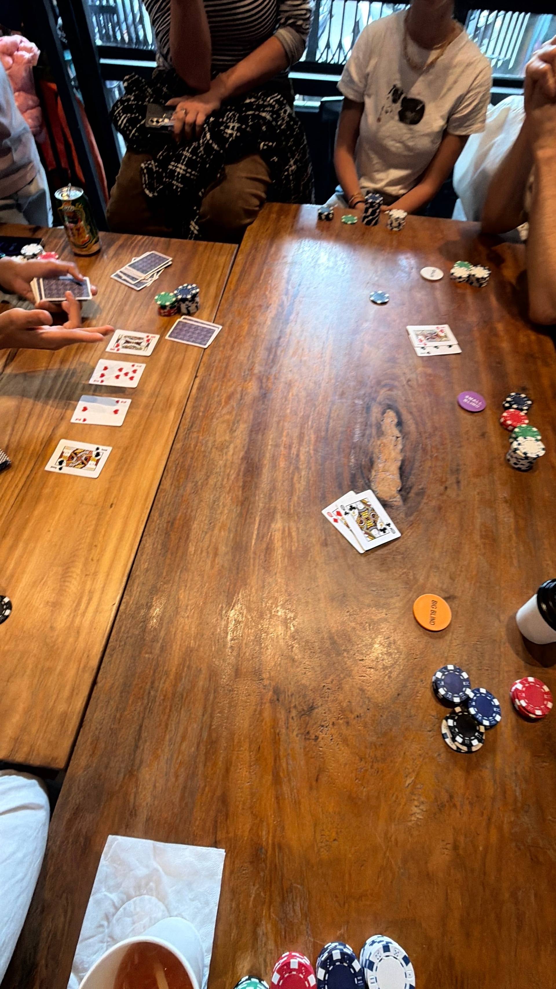 A lively poker tournament scene unfolds with multiple players gathered around wooden tables in a well-lit Đà Lạt gaming venue. The relaxed, social atmosphere captures friends enjoying card games together.