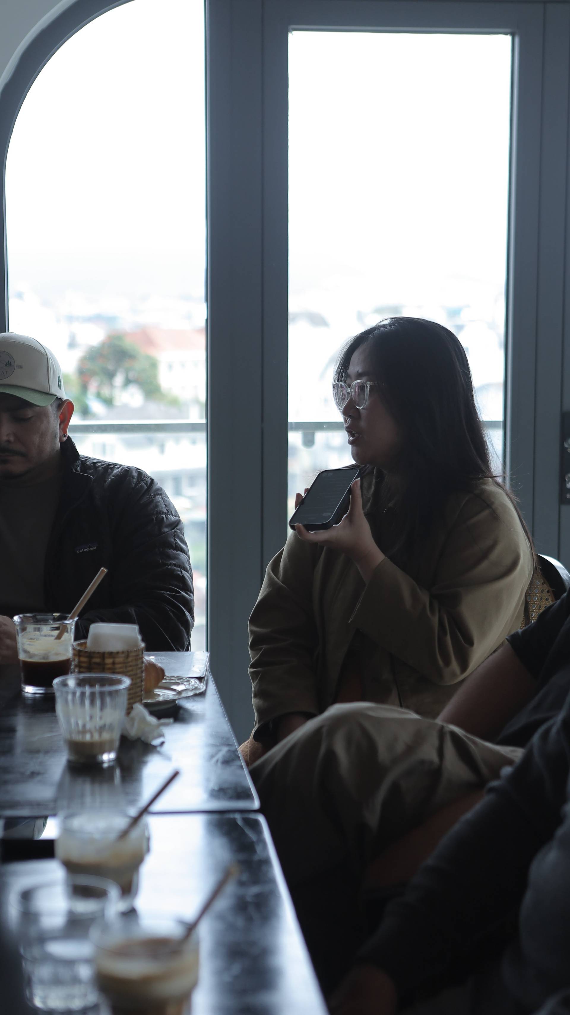 A group of friends enjoys a casual conversation over drinks in a cozy café with large windows overlooking Đà Lạt's scenic landscape.