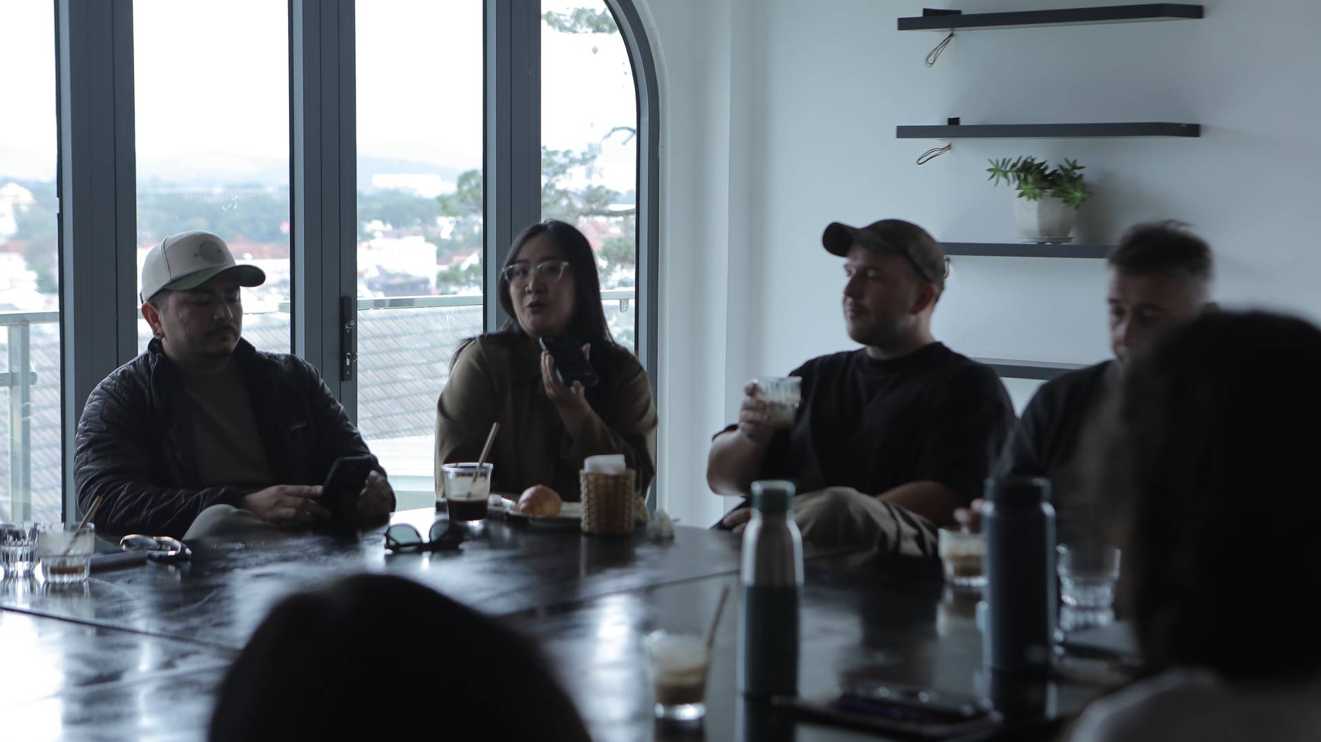 A group of friends enjoys a casual gathering in a cozy café with large windows overlooking Đà Lạt's scenic landscape.