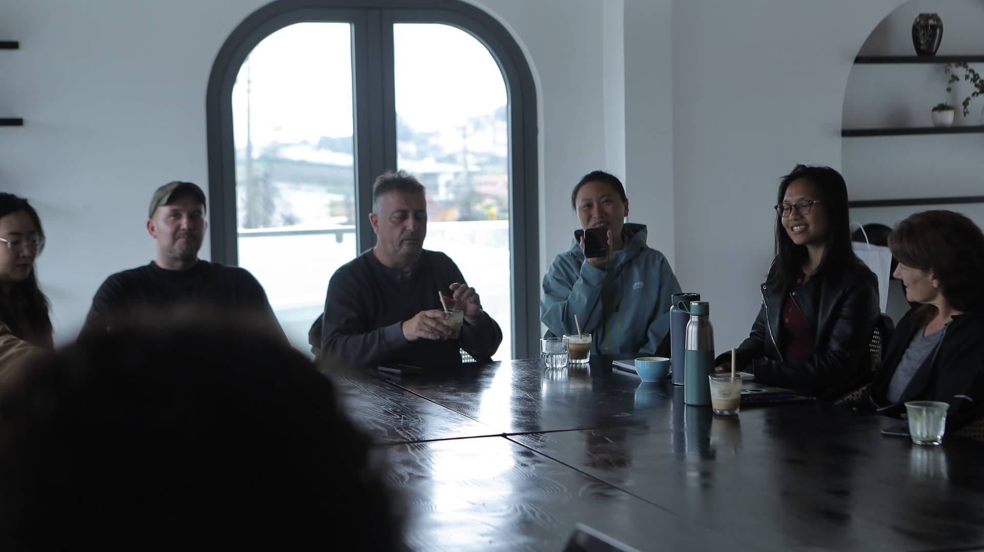 A group of friends gathered around a long table in Đà Lạt, Vietnam, enjoying drinks and engaging in conversation with scenic views through large windows.