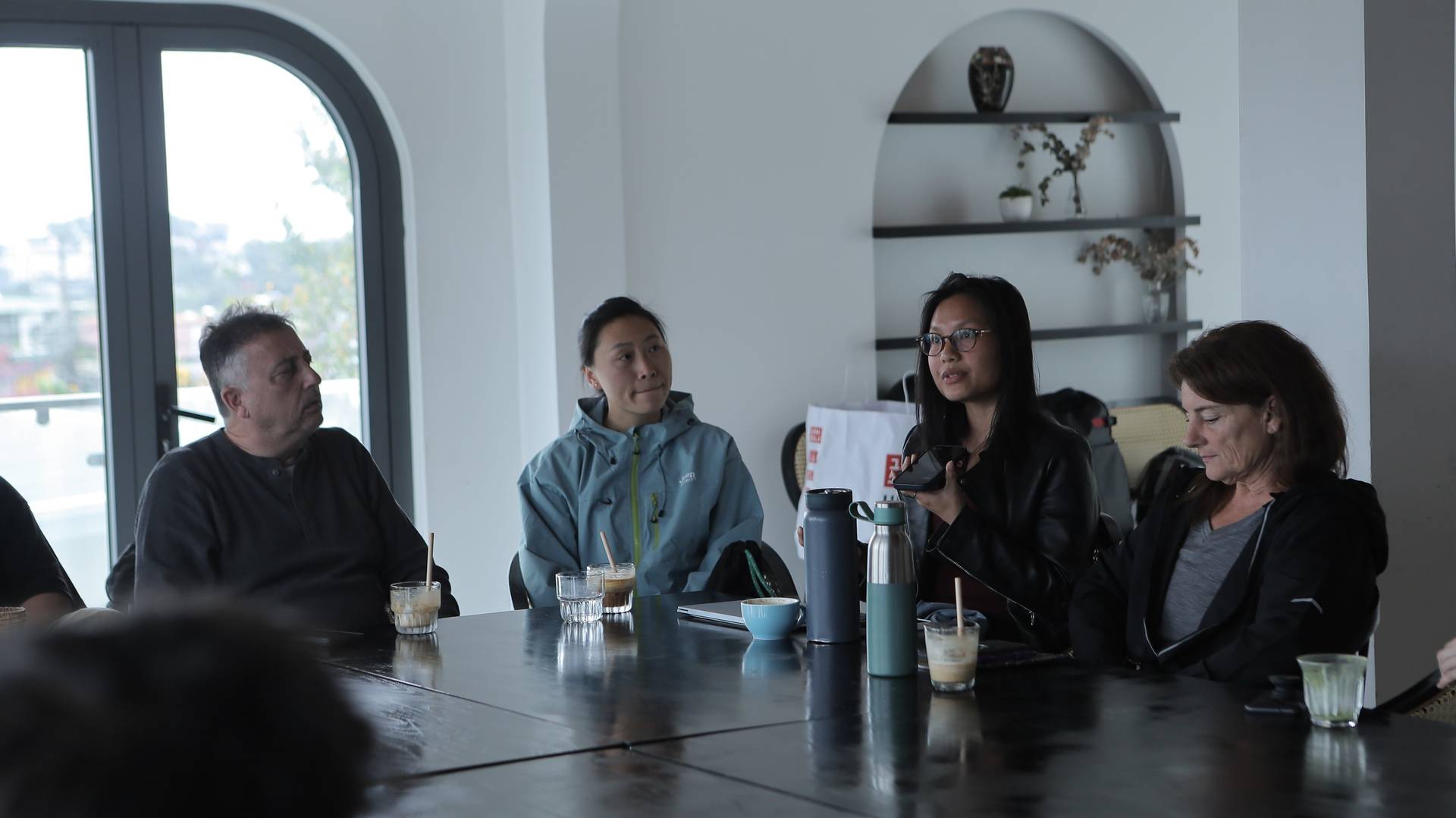 A group of people engaged in a casual meeting or discussion around a table in a modern café setting with natural light streaming through large windows.