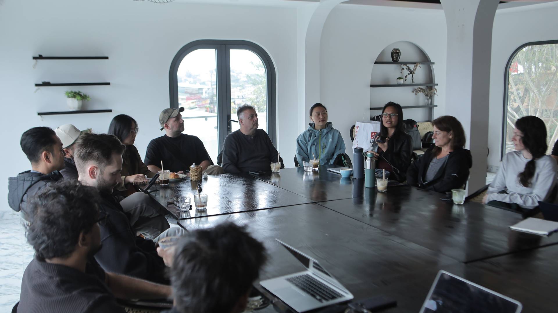 A group of people engaged in a discussion around a long table in a modern café setting with large windows overlooking Đà Lạt's scenic landscape.
