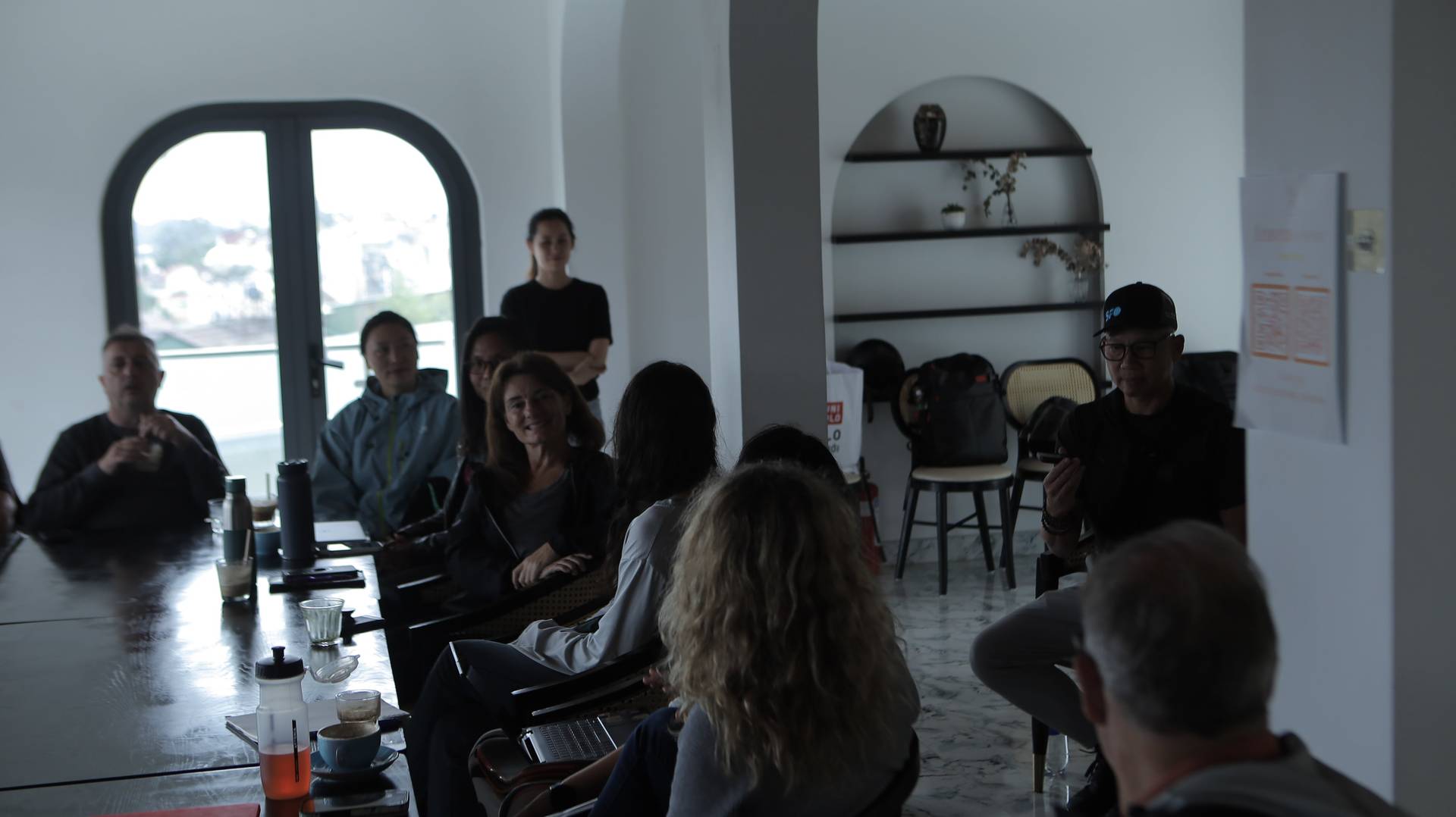 A group of people is gathered in a modern, well-lit room with large windows overlooking Đà Lạt's scenic landscape, engaging in what appears to be an informal meeting or workshop led by someone seated at the front.