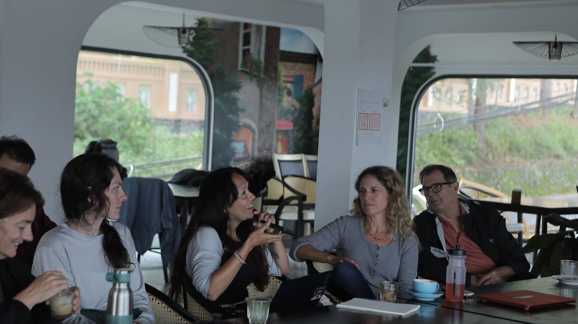 A group of people enjoying a casual conversation in a cozy café with scenic views of Đà Lạt's charming architecture and greenery.