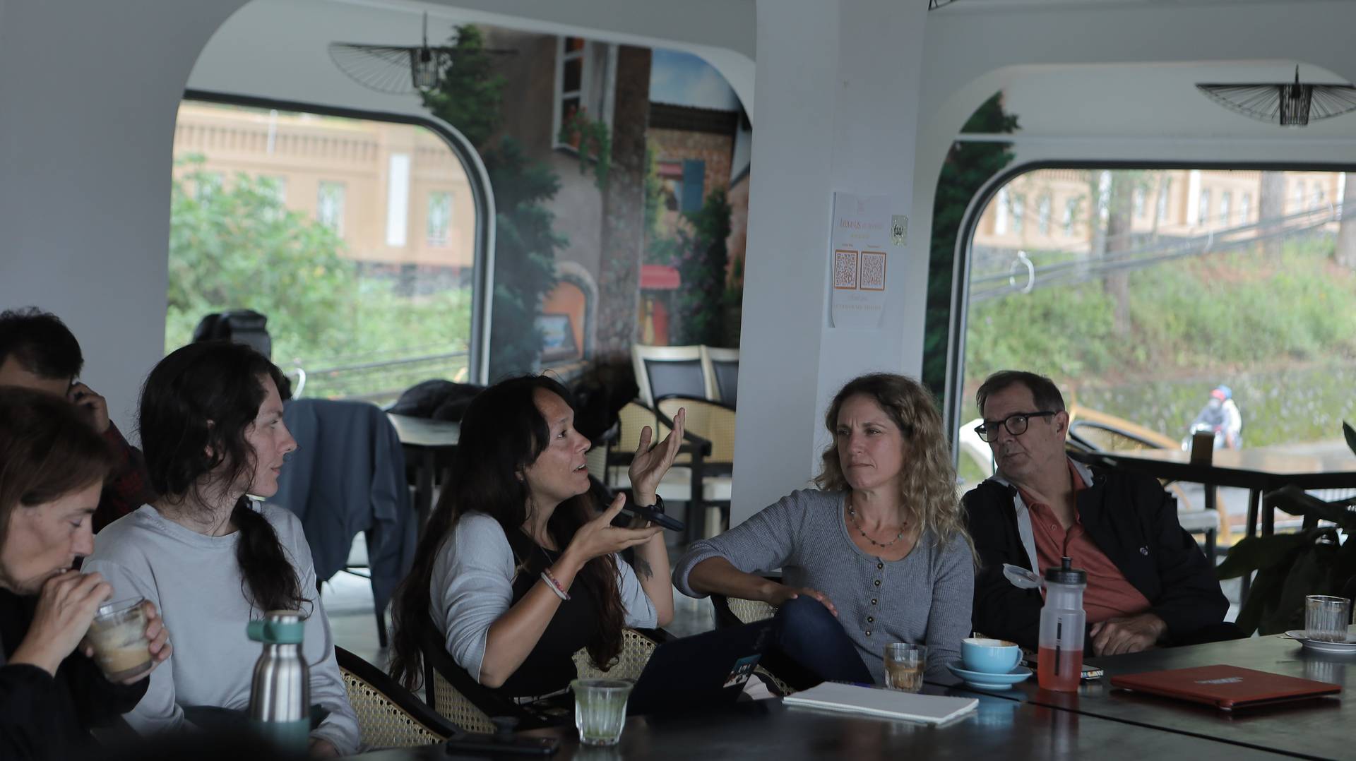 A group of people engaged in conversation over drinks at a café in Đà Lạt, Vietnam, with scenic views visible through large windows.