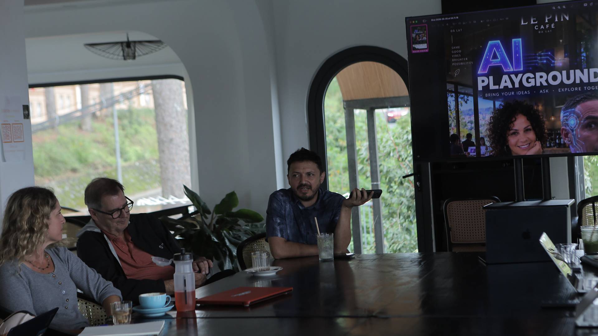 A group of people engaged in a discussion over drinks and laptops in a cozy café setting with scenic views of Đà Lạt.