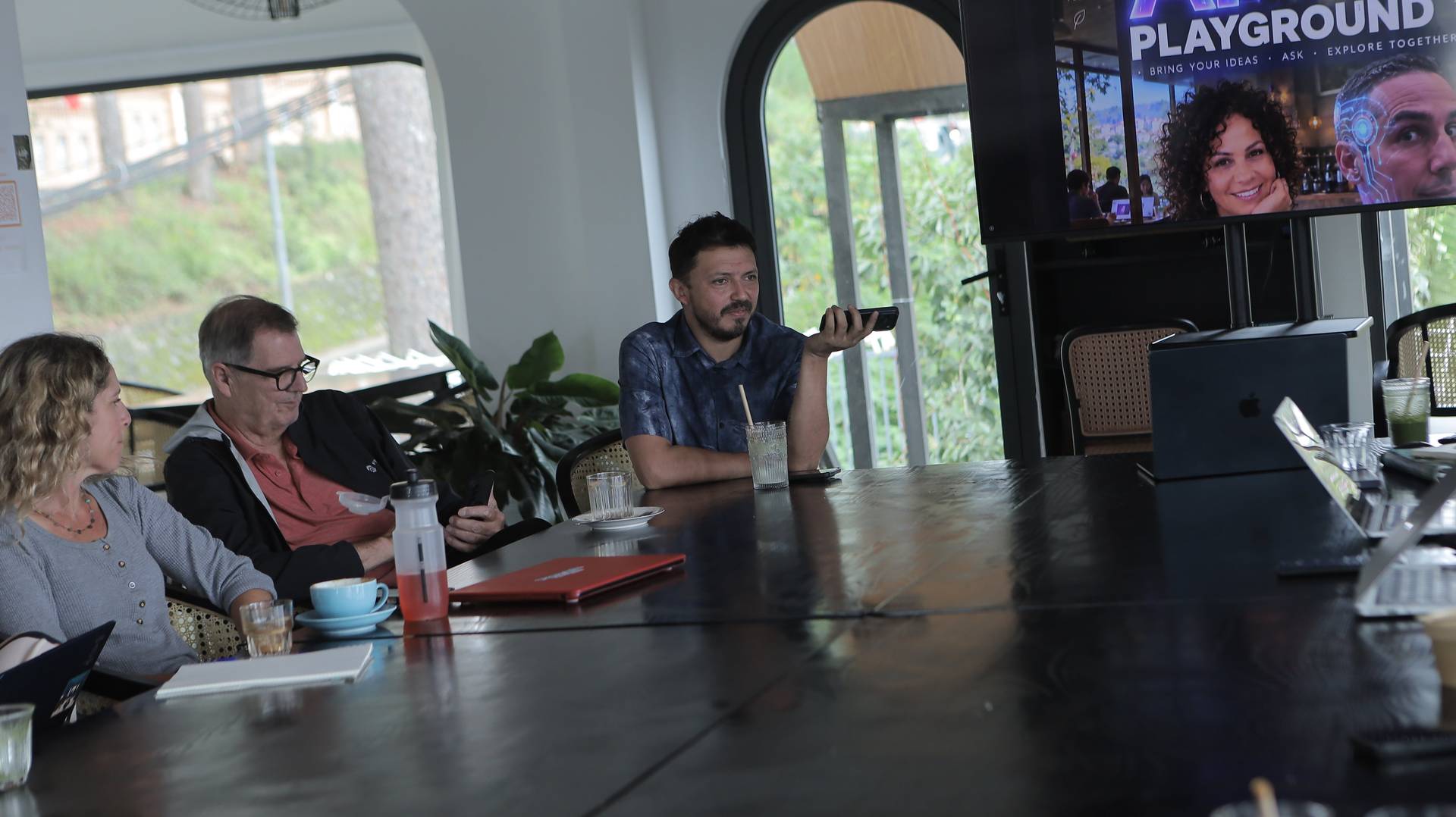 A group of people engaged in a discussion around a table in a modern café setting with large windows overlooking greenery, suggesting a collaborative or brainstorming session.