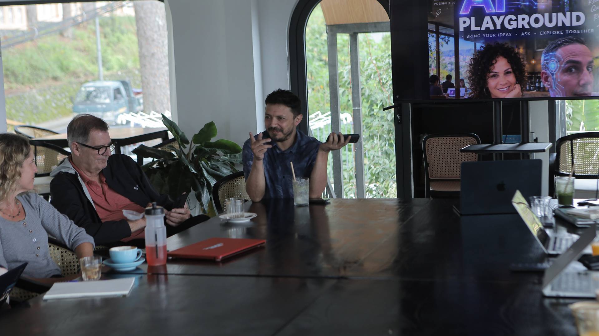 A group of people engaged in a discussion around a table in a modern café setting with large windows overlooking greenery and a sign that reads 'AI PLAYGROUND'.