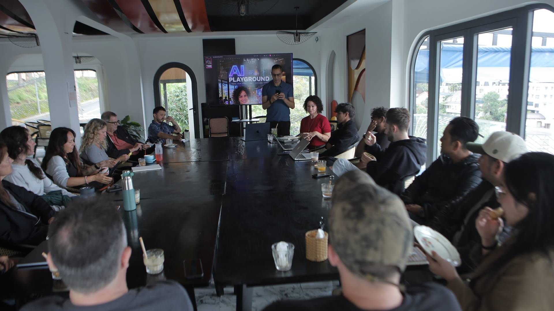 A group of people gathered around a large table in a modern café setting in Đà Lạt, Vietnam, participating in an AI-themed workshop or discussion.