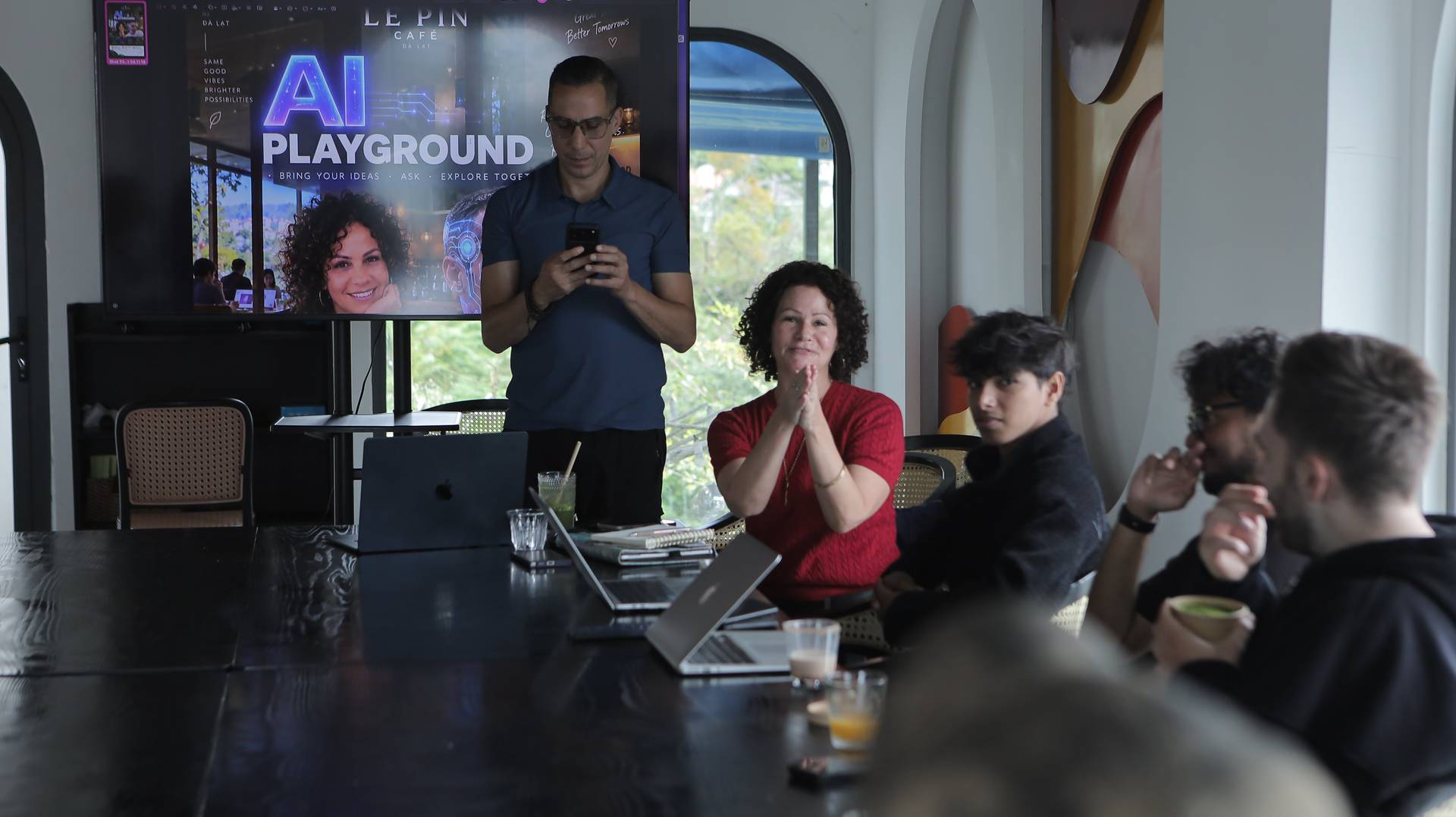 A group of professionals is gathered in a modern café in Đà Lạt, Vietnam, participating in an AI workshop titled 'AI Playground'. They are engaged in discussion and collaboration around laptops on a sleek black table.