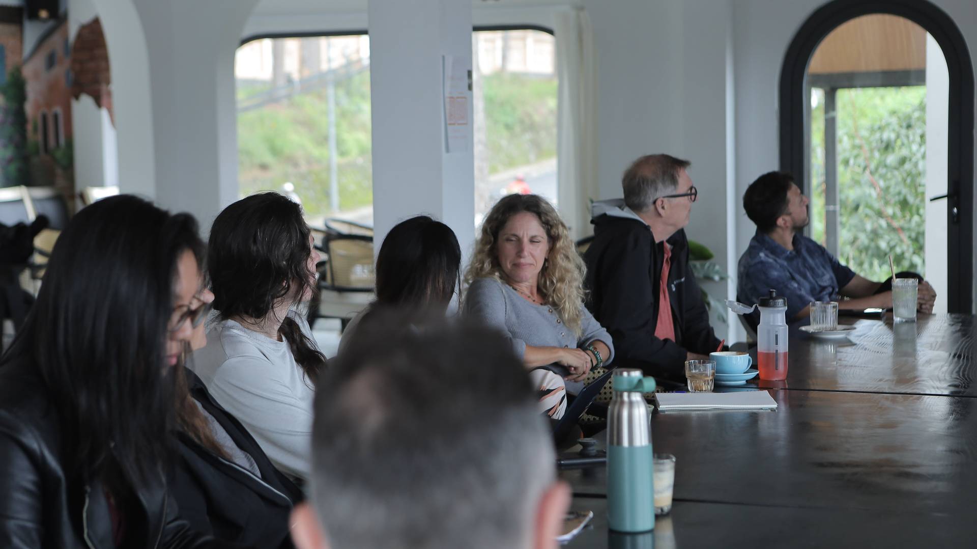A group of people engaged in a discussion around a long table in a modern café setting with large windows overlooking greenery.