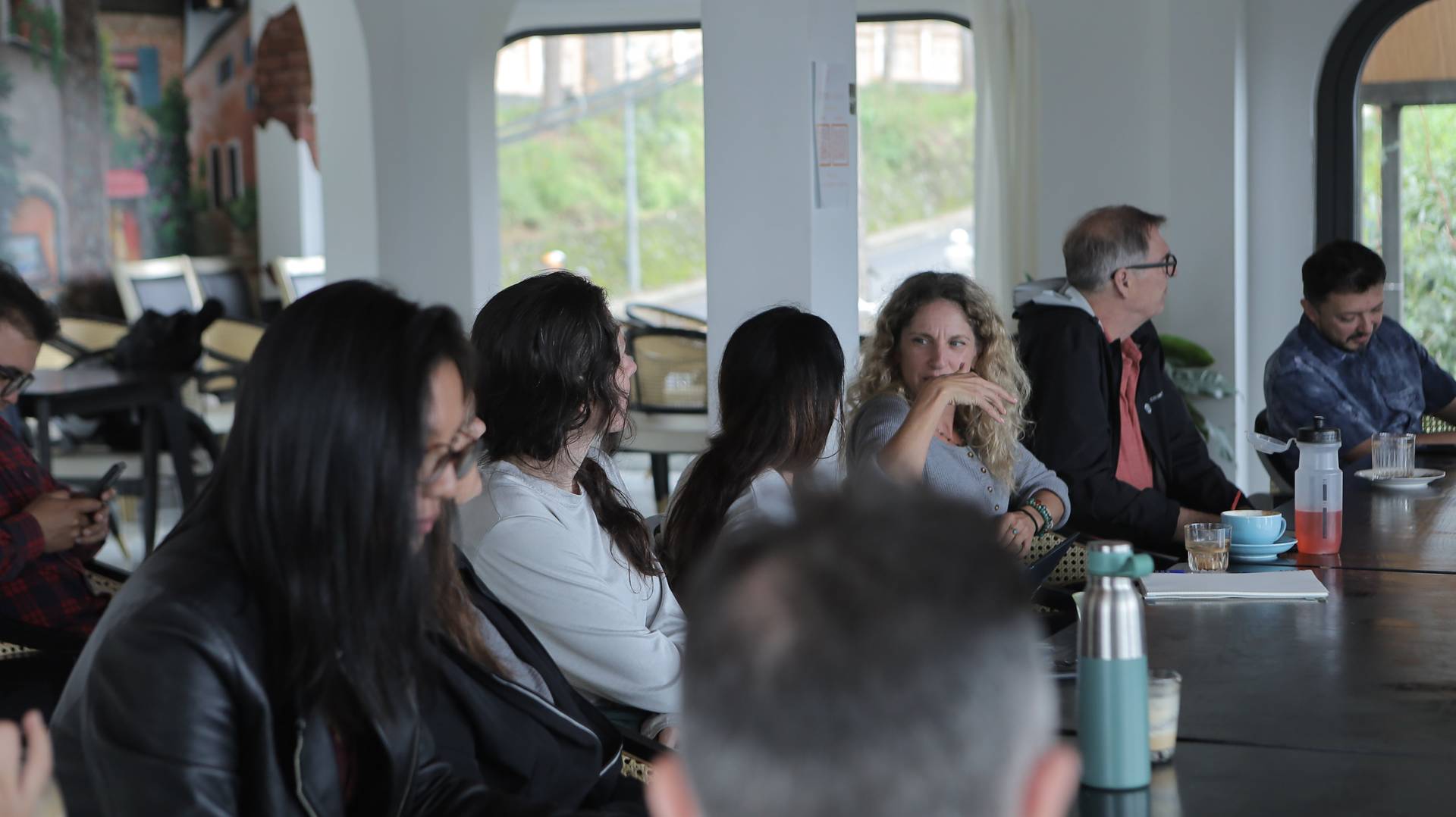 A group of people engaged in a discussion around a long table in a modern café setting with large windows overlooking greenery.