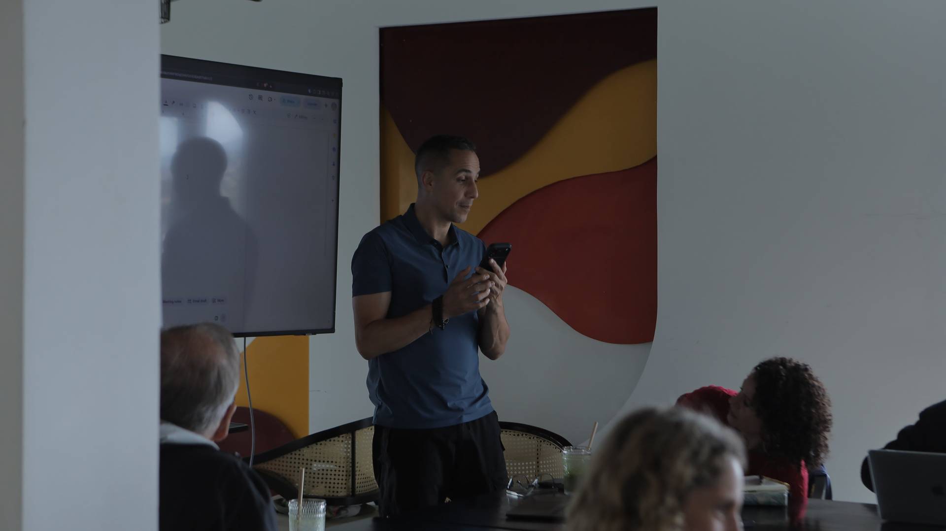 A man presents to a small audience in a modern setting with vibrant wall art and a projection screen displaying digital content.